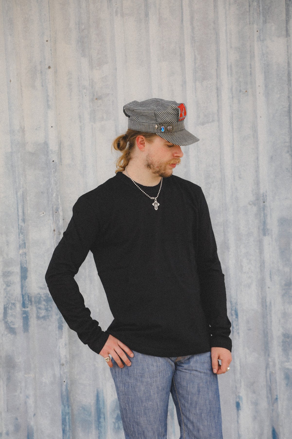 A man wearing a black long-sleeve shirt, blue jeans, a gray cap, and a silver cross necklace stands against a textured gray wall.