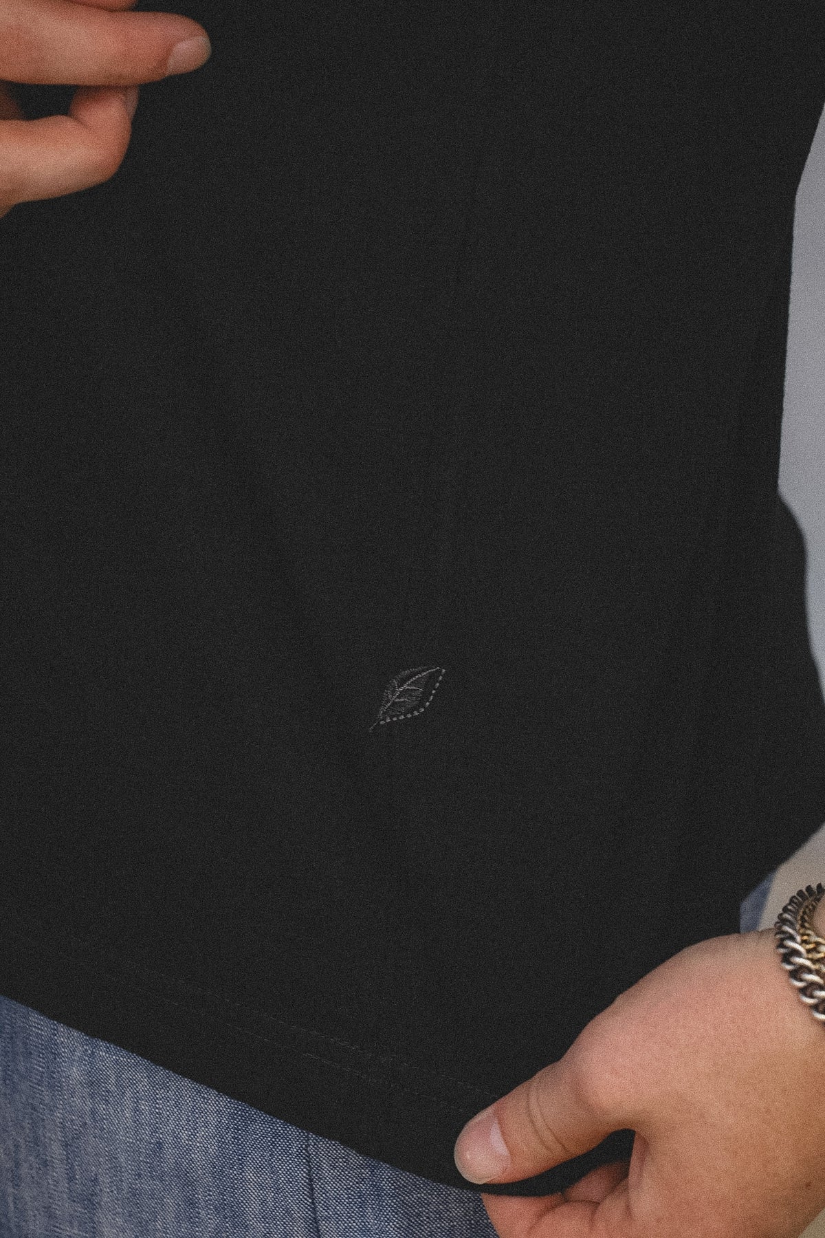 Close-up of a person wearing a black t-shirt with a small embroidered logo on the front, paired with blue jeans and a gold chain bracelet.