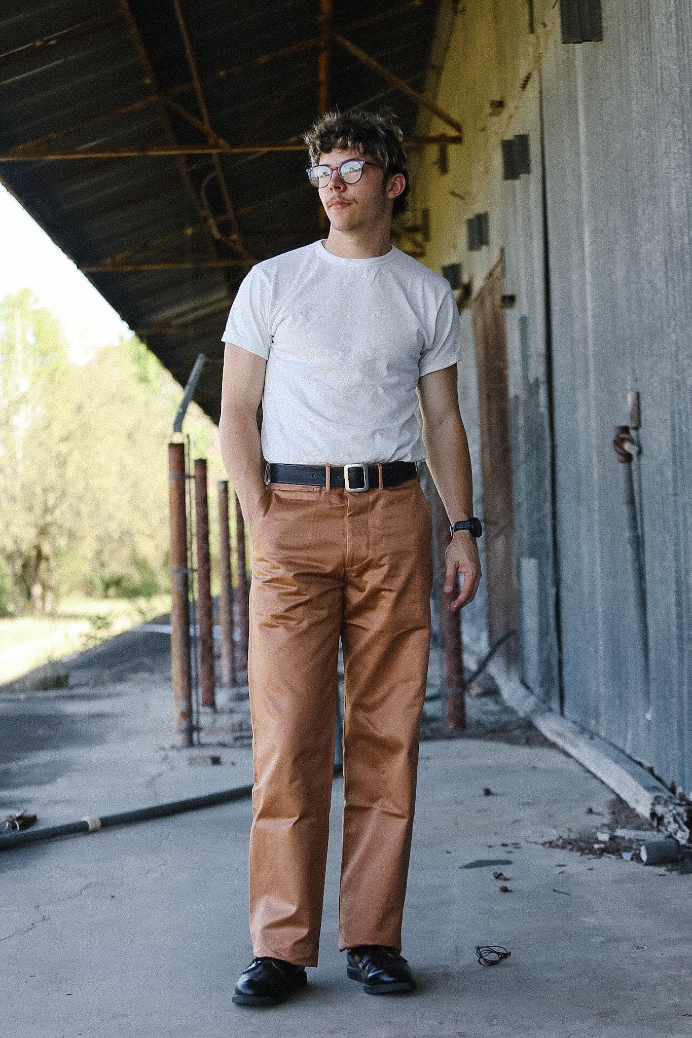 Man wearing a white t-shirt, brown pants, and glasses standing in front of a building.