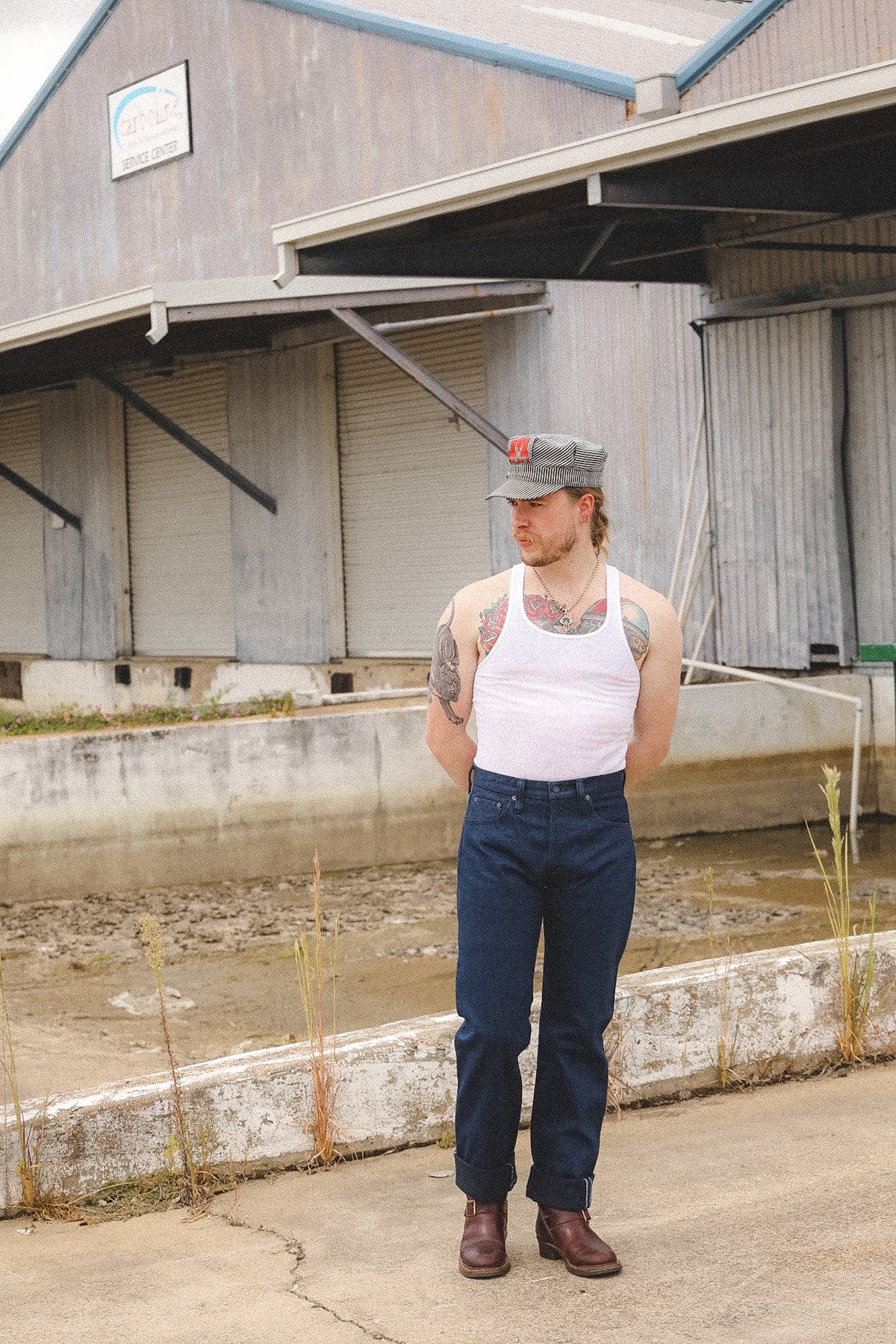 A man stands outdoors in front of an industrial building, wearing a white tank top, dark blue jeans, brown boots, and a gray baseball cap.