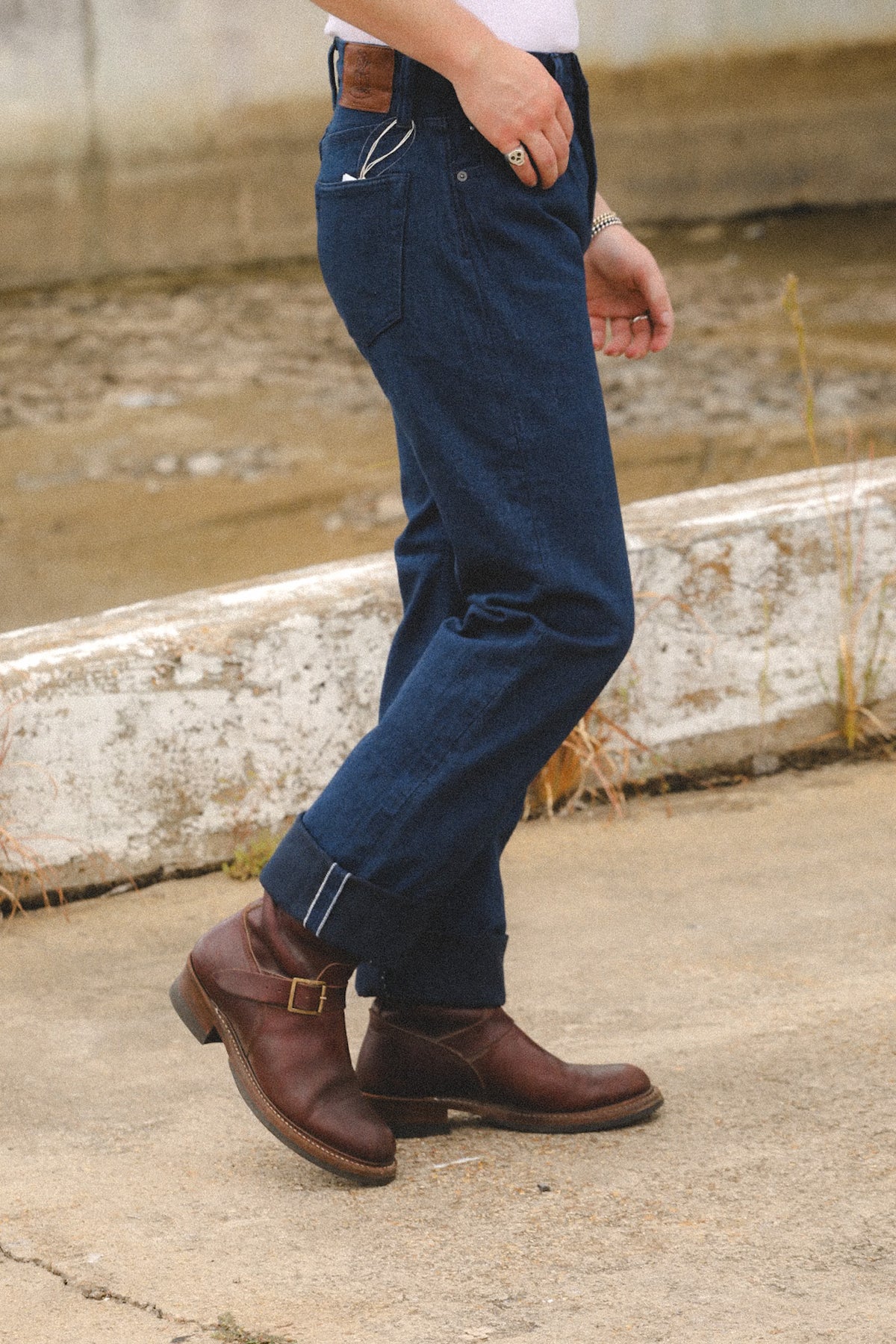 A person wearing dark blue denim jeans with rolled cuffs and brown leather boots, standing outdoors on a concrete surface near a weathered white wall.
