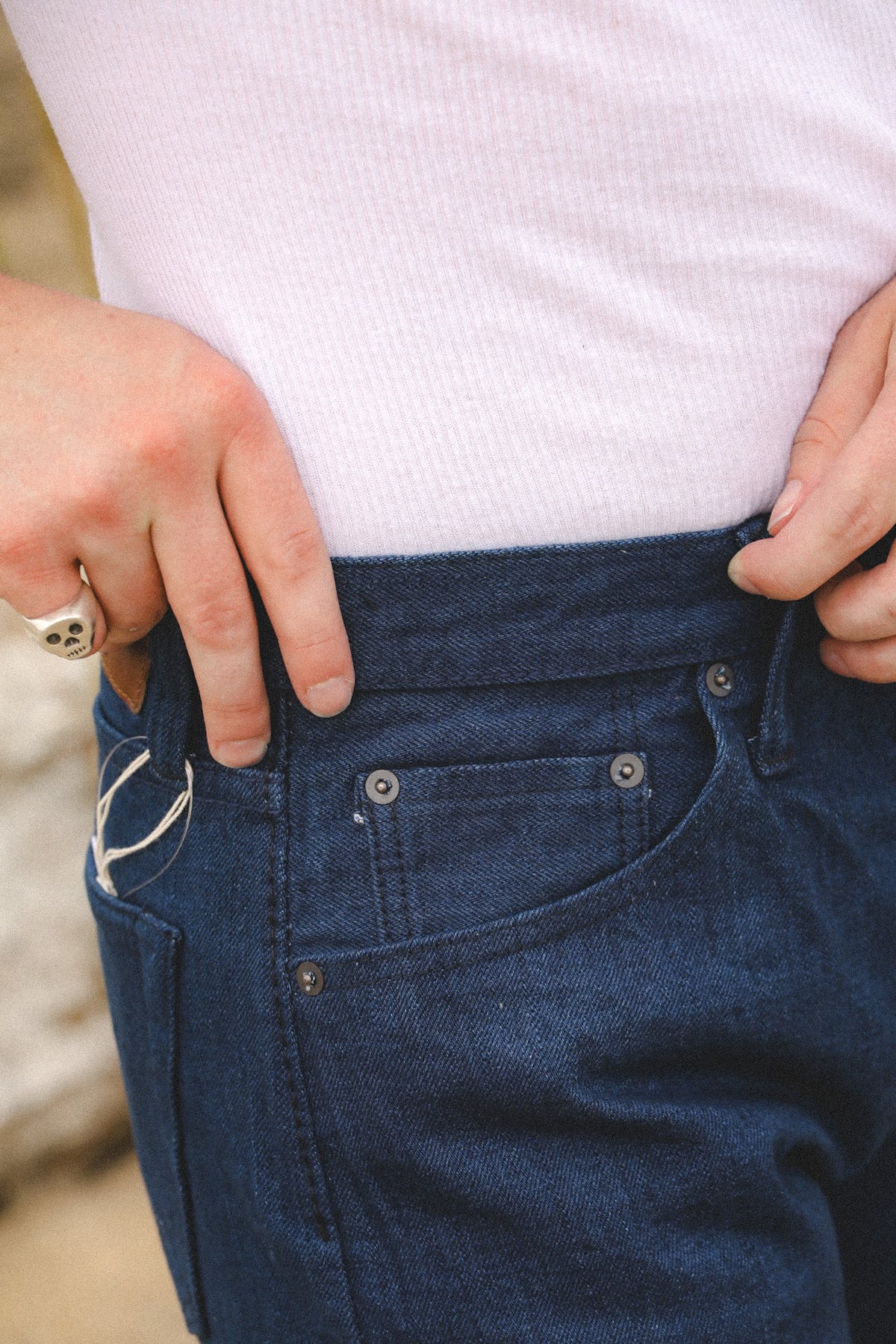 Close-up of a person wearing dark blue denim jeans and a light pink ribbed top, pulling the waistband of the jeans slightly outward with both hands.