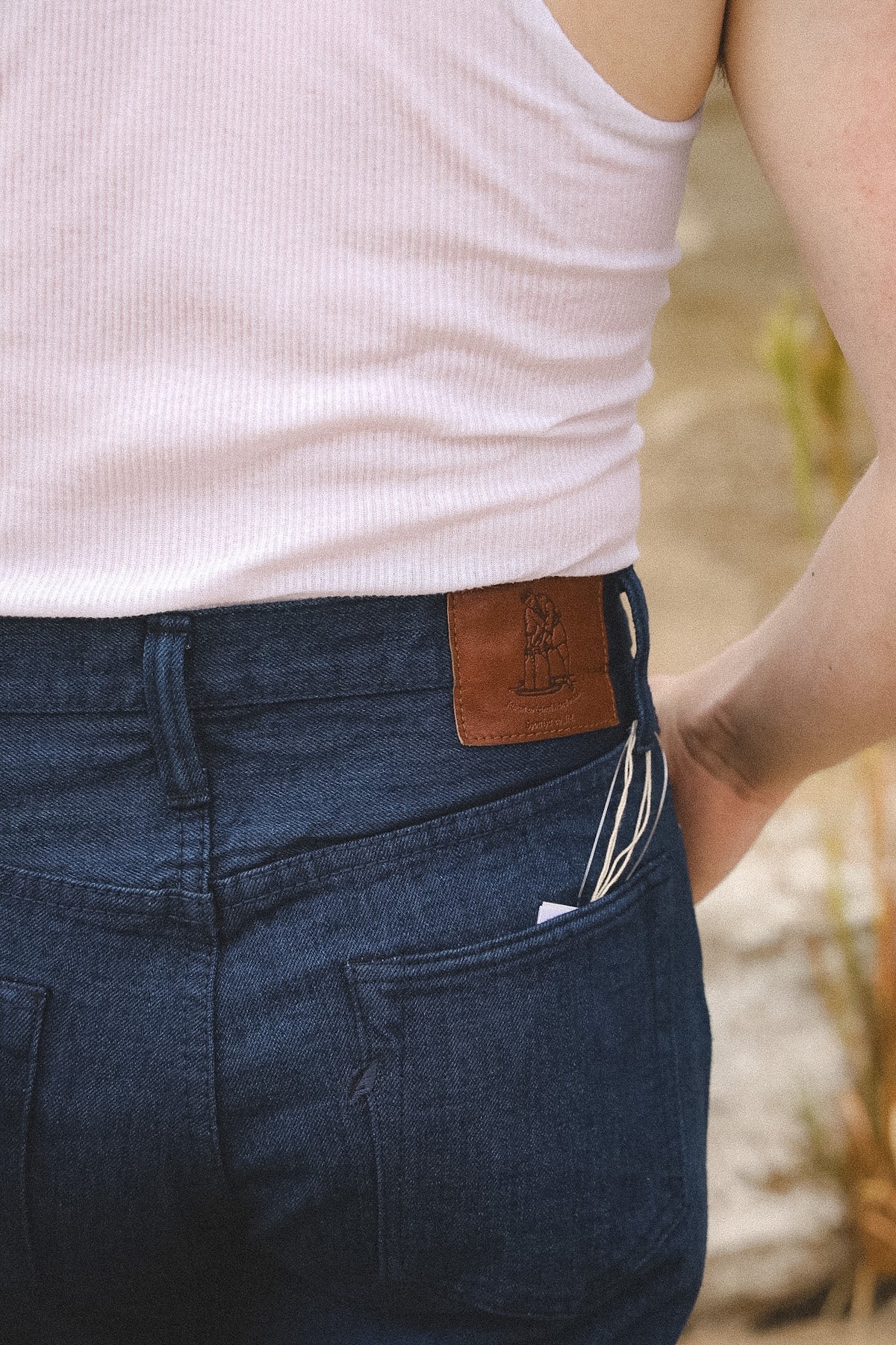 Close-up of the back waist of a person wearing dark blue denim jeans and a white ribbed tank top, with a brown leather patch on the back waistband and a phone tucked into the pocket.