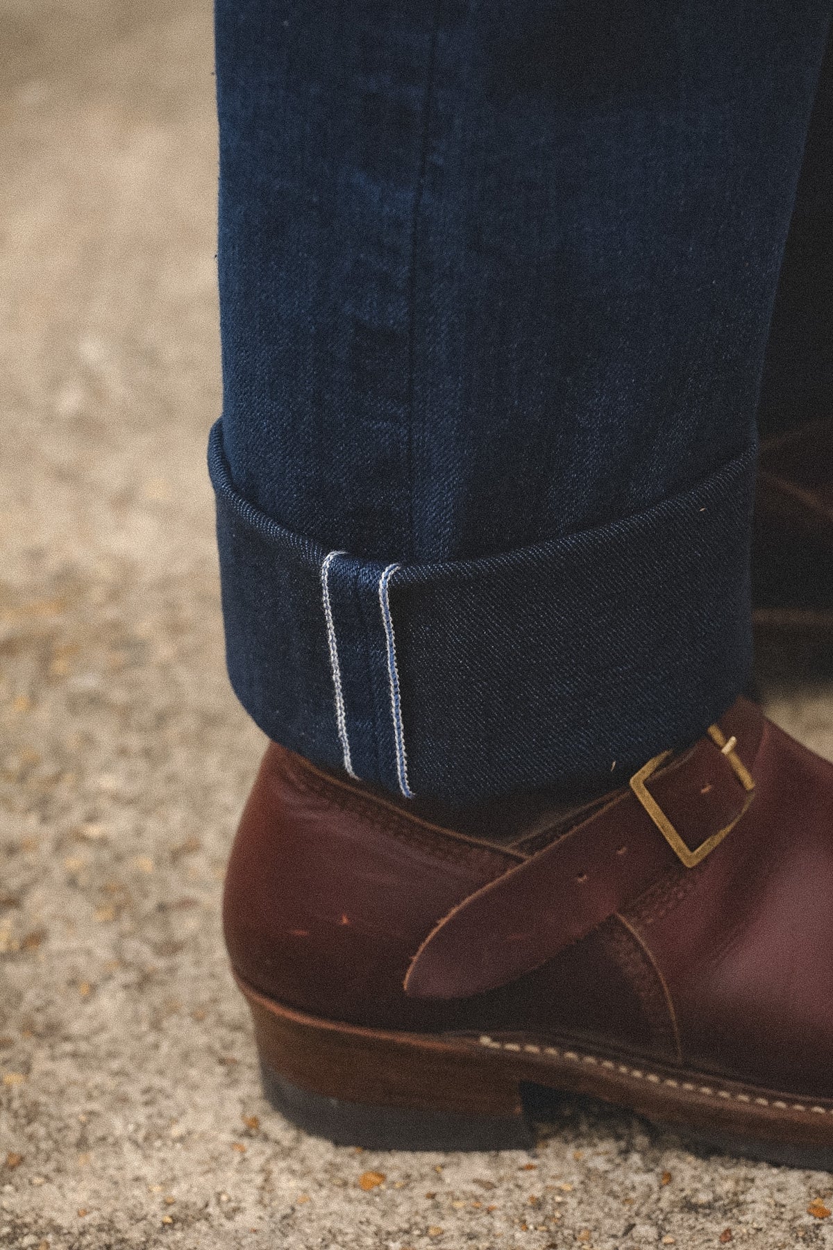Close-up of a person's lower leg wearing dark blue denim jeans with a rolled cuff showing white contrast stitching, paired with a brown leather buckle shoe on a textured concrete ground.