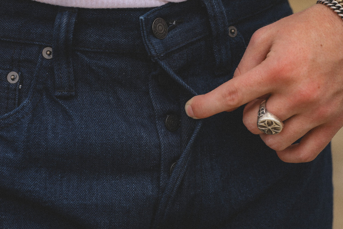 Close-up of a person wearing dark indigo denim jeans, pointing at the button area with their right hand. The person is also wearing a silver ring and a chain bracelet.