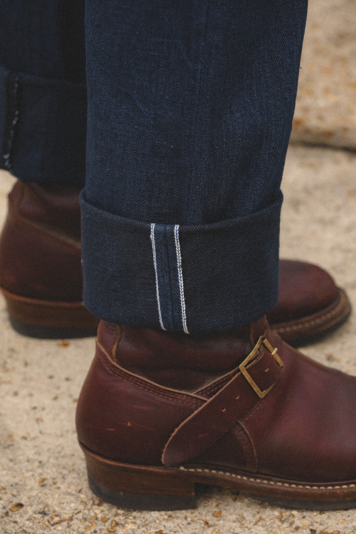 Close-up of a person's legs wearing dark blue denim jeans with a rolled cuff showing double white stitching, paired with dark brown leather buckle-strap boots on a light concrete surface.