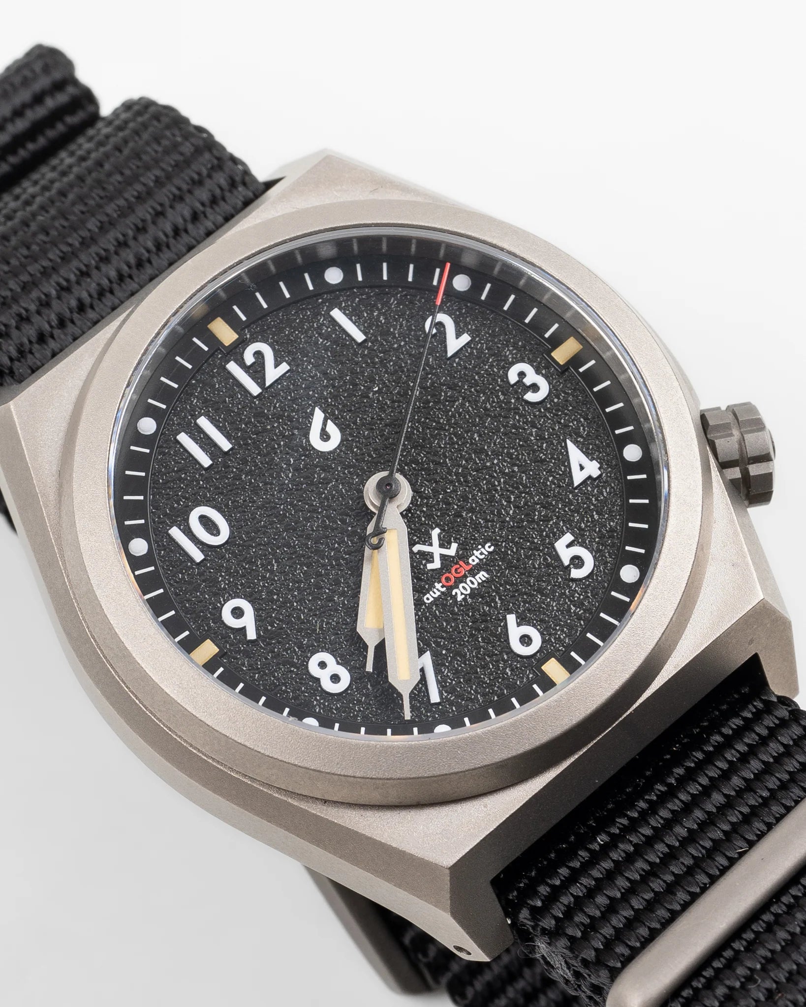 Close-up of OGL autoGLatic Bold MK1 field watch with black textured dial, lumed hands, beige accents, and black nylon strap