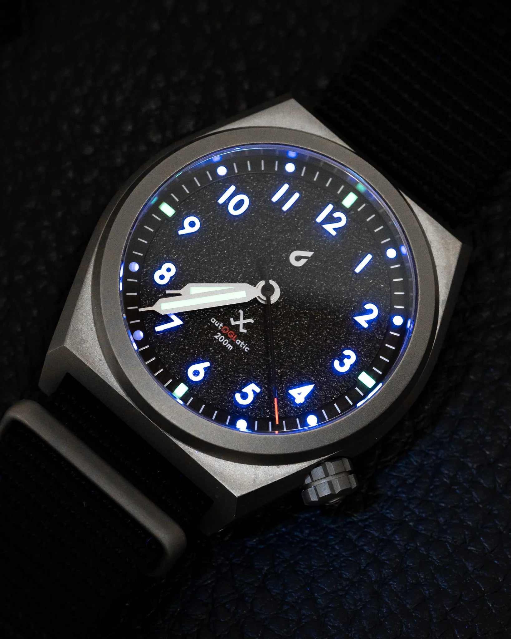 Close-up of autOGLatic Field Watch with luminous blue numerals and green accents on black face, black nylon strap