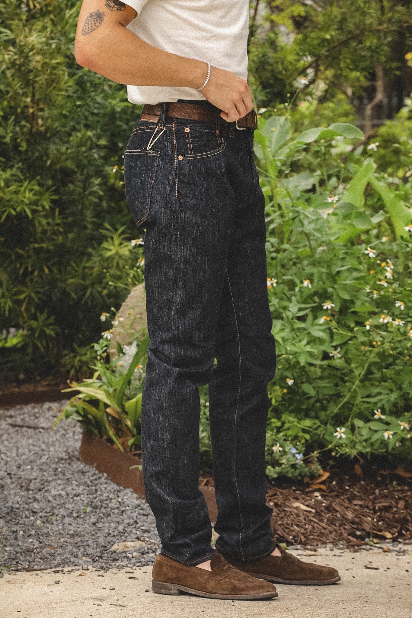 A person wearing dark indigo denim jeans, a white t-shirt, and brown suede loafers, standing outdoors in front of green garden foliage.
