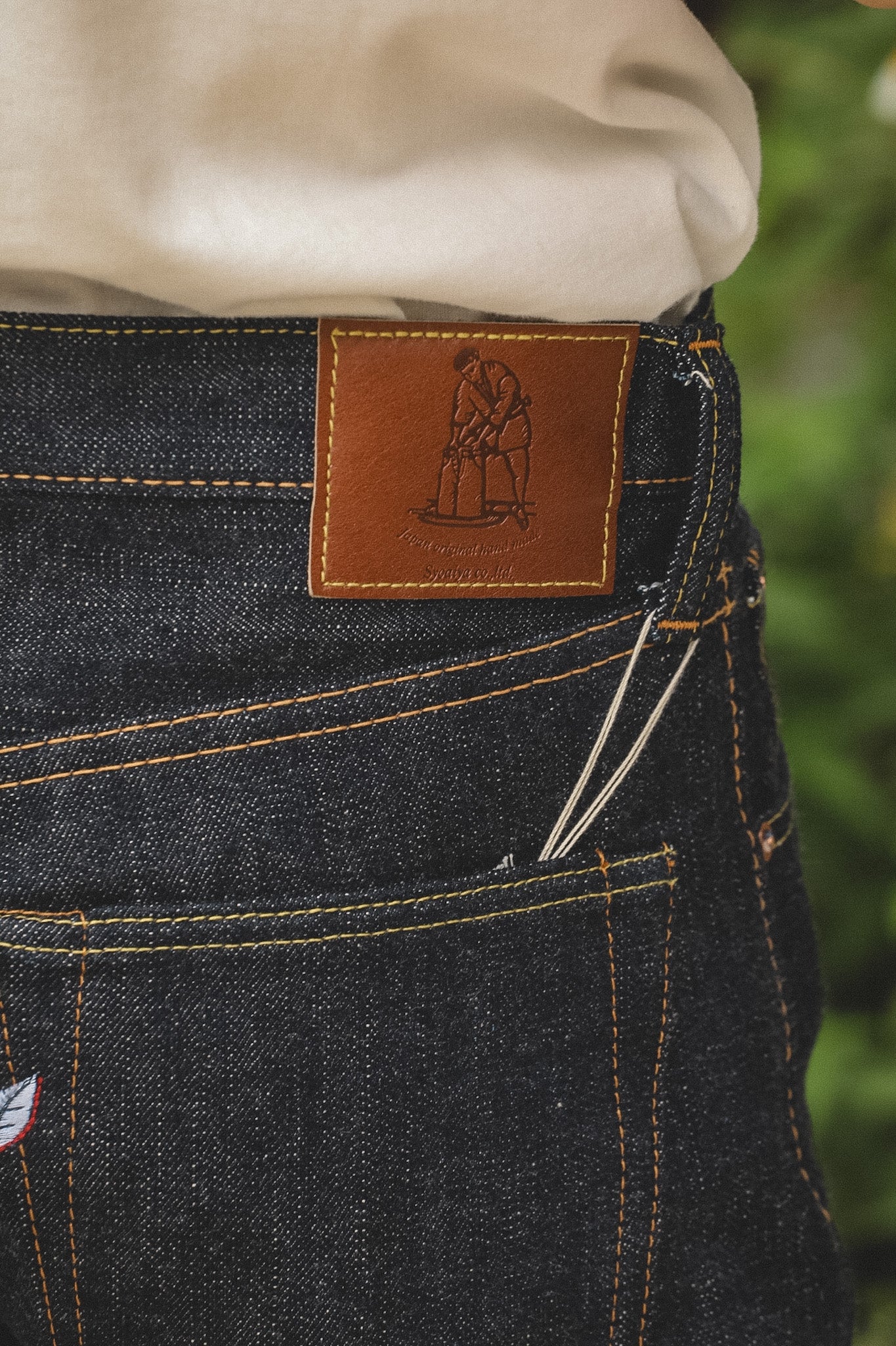 Close-up of the back waistband of dark indigo denim jeans with a brown leather patch showing the 'Hagasa' brand logo, worn with a cream-colored shirt.