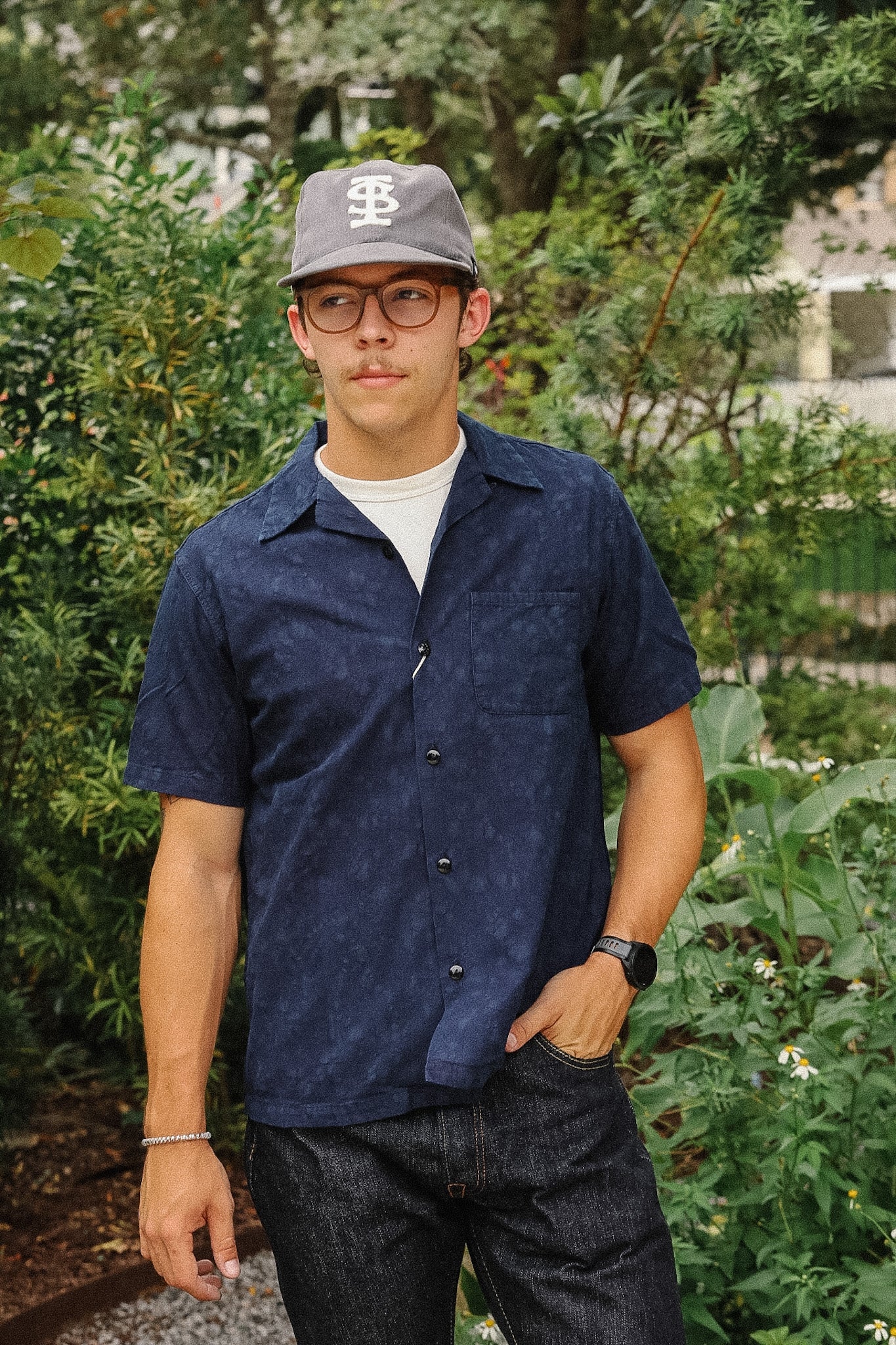 A man wearing a navy blue short-sleeve button-up shirt, dark jeans, a gray baseball cap with a white 'BS' logo, and glasses, standing outdoors in front of green foliage.