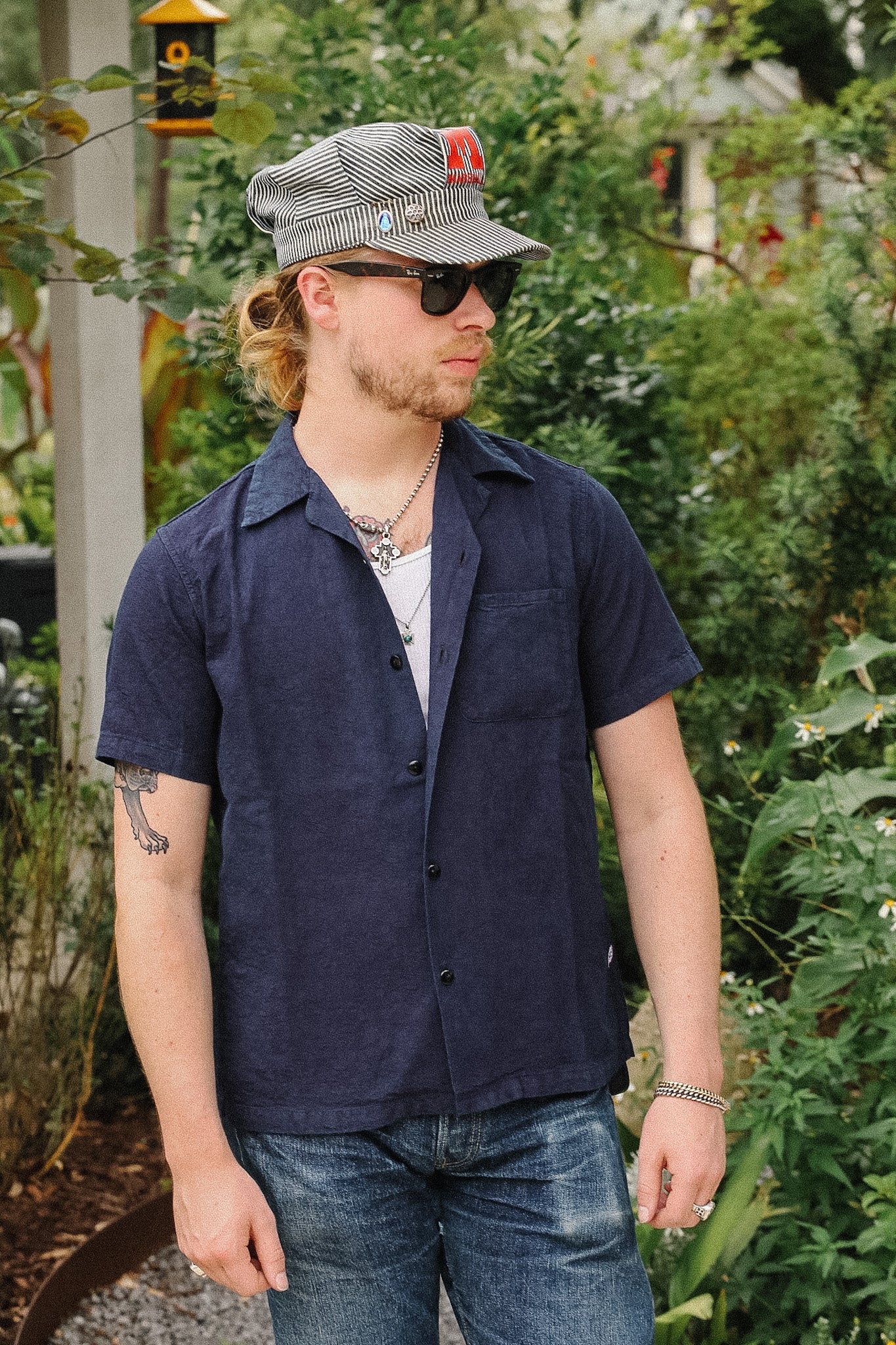 A man wearing a navy blue short-sleeve button-up shirt, blue jeans, a gray patterned cap, and sunglasses, standing outdoors in a garden setting.
