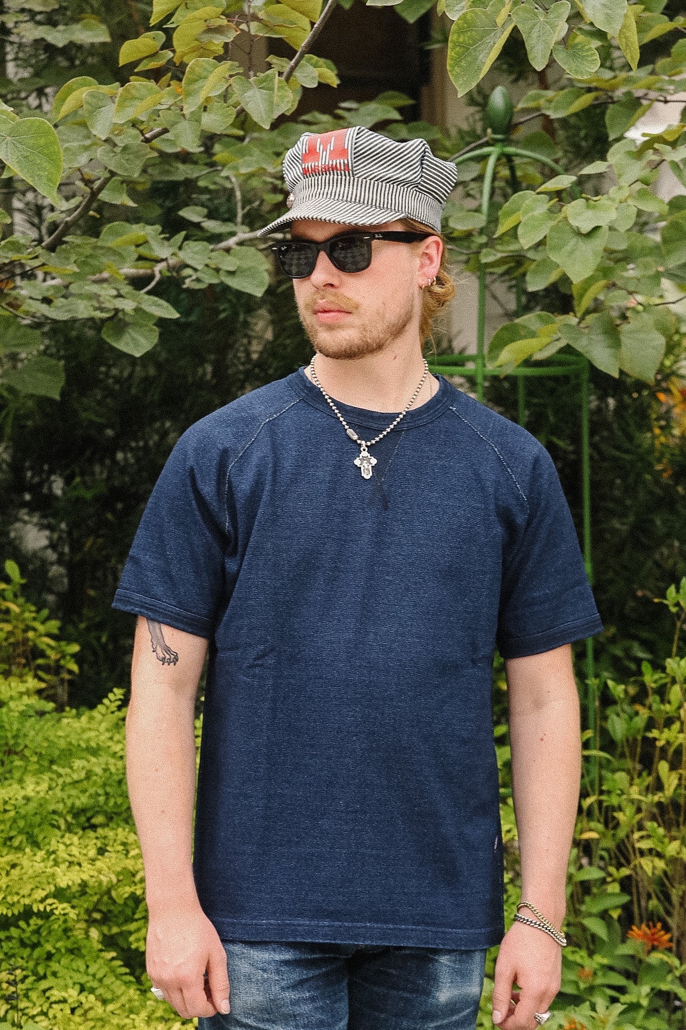 A man wearing a dark blue short-sleeve raglan t-shirt, a striped baseball cap, and sunglasses, standing outdoors in front of green foliage.
