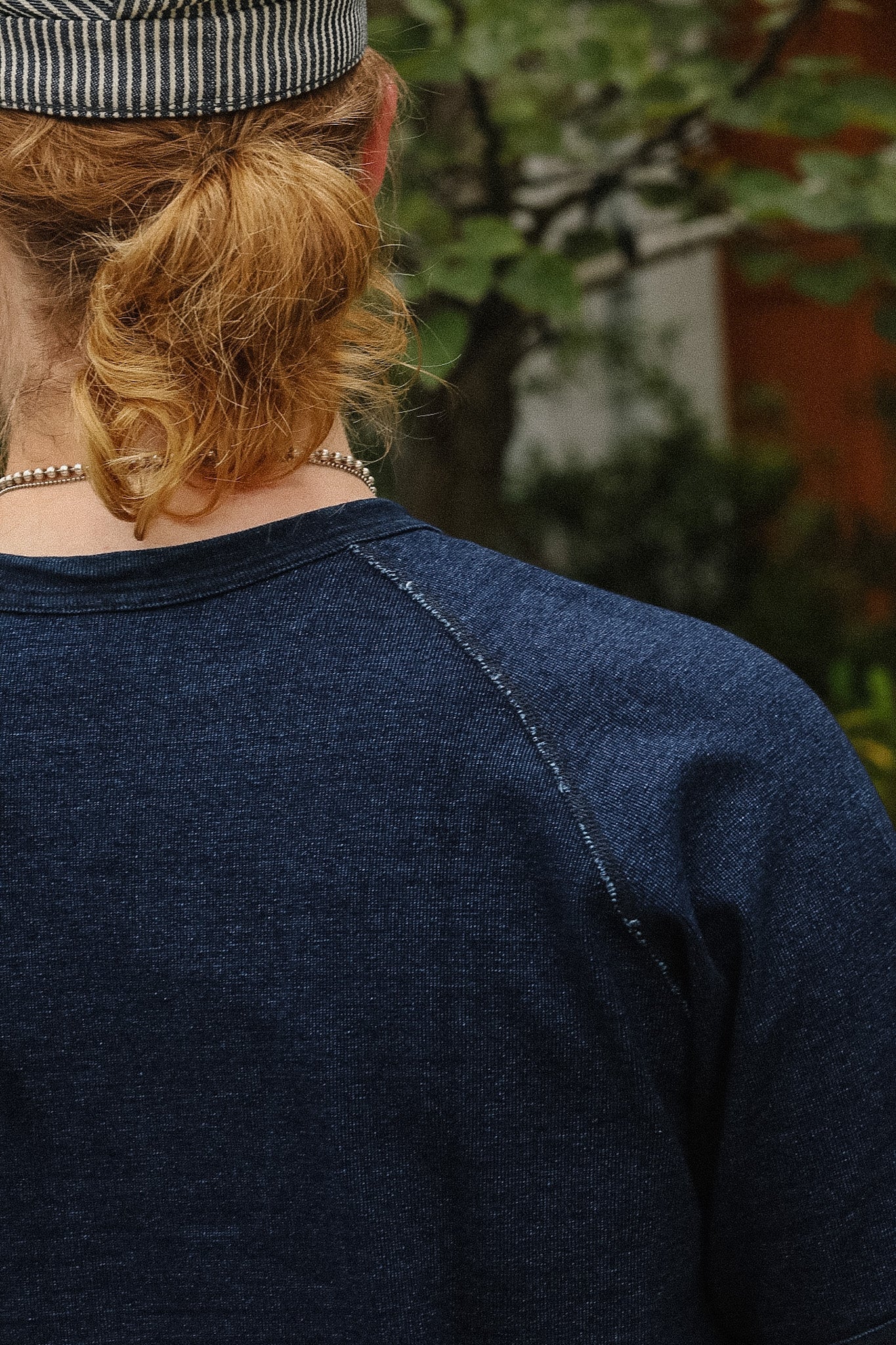 Back view of a person wearing a dark navy blue heathered long-sleeve raglan shirt, with a striped cap and pearl necklace visible, set against a blurred outdoor background.