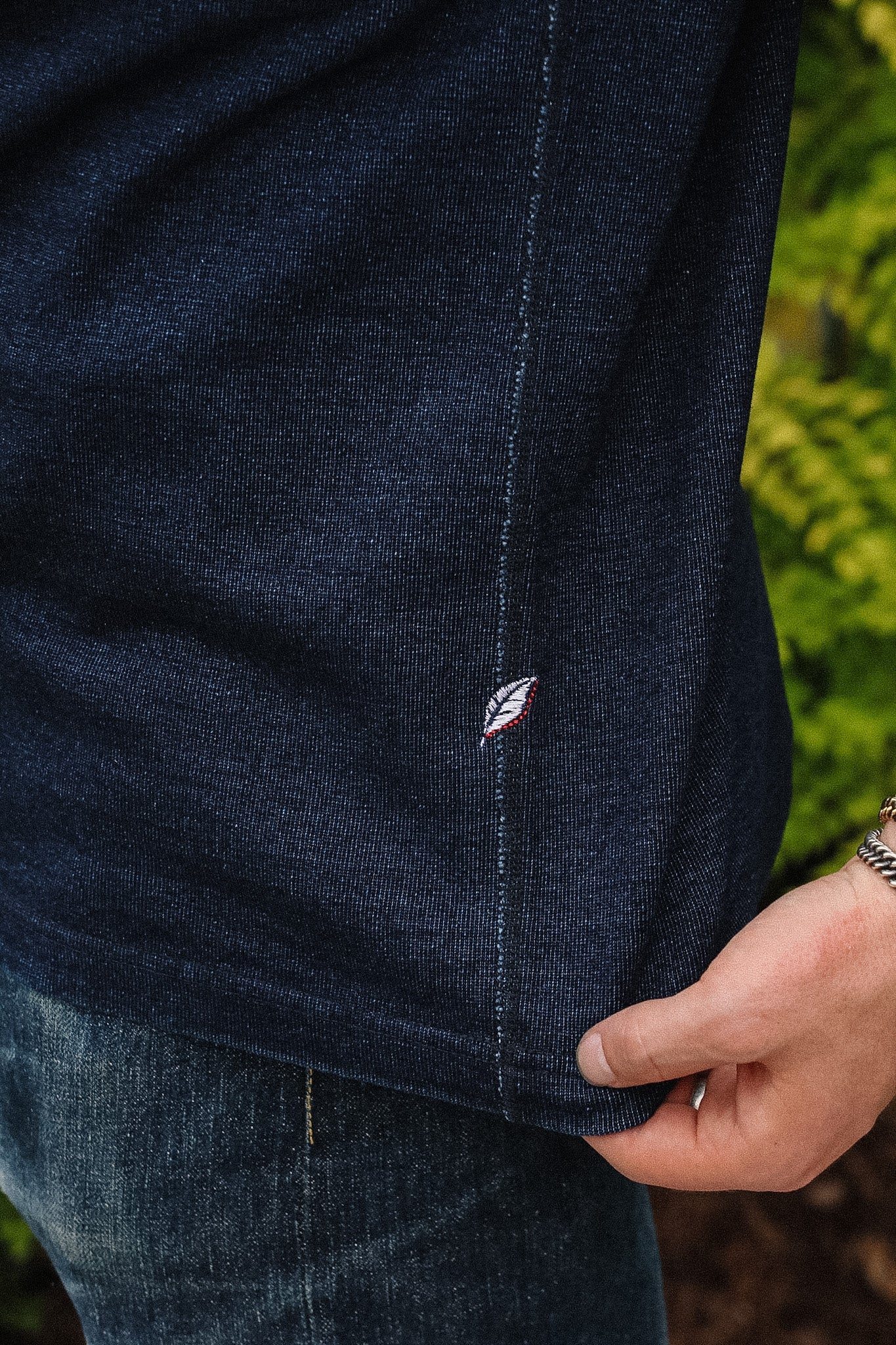 Close-up of a person wearing a dark navy blue ribbed knit top, pulling the hem slightly to show the texture and a small embroidered logo, with green foliage blurred in the background.