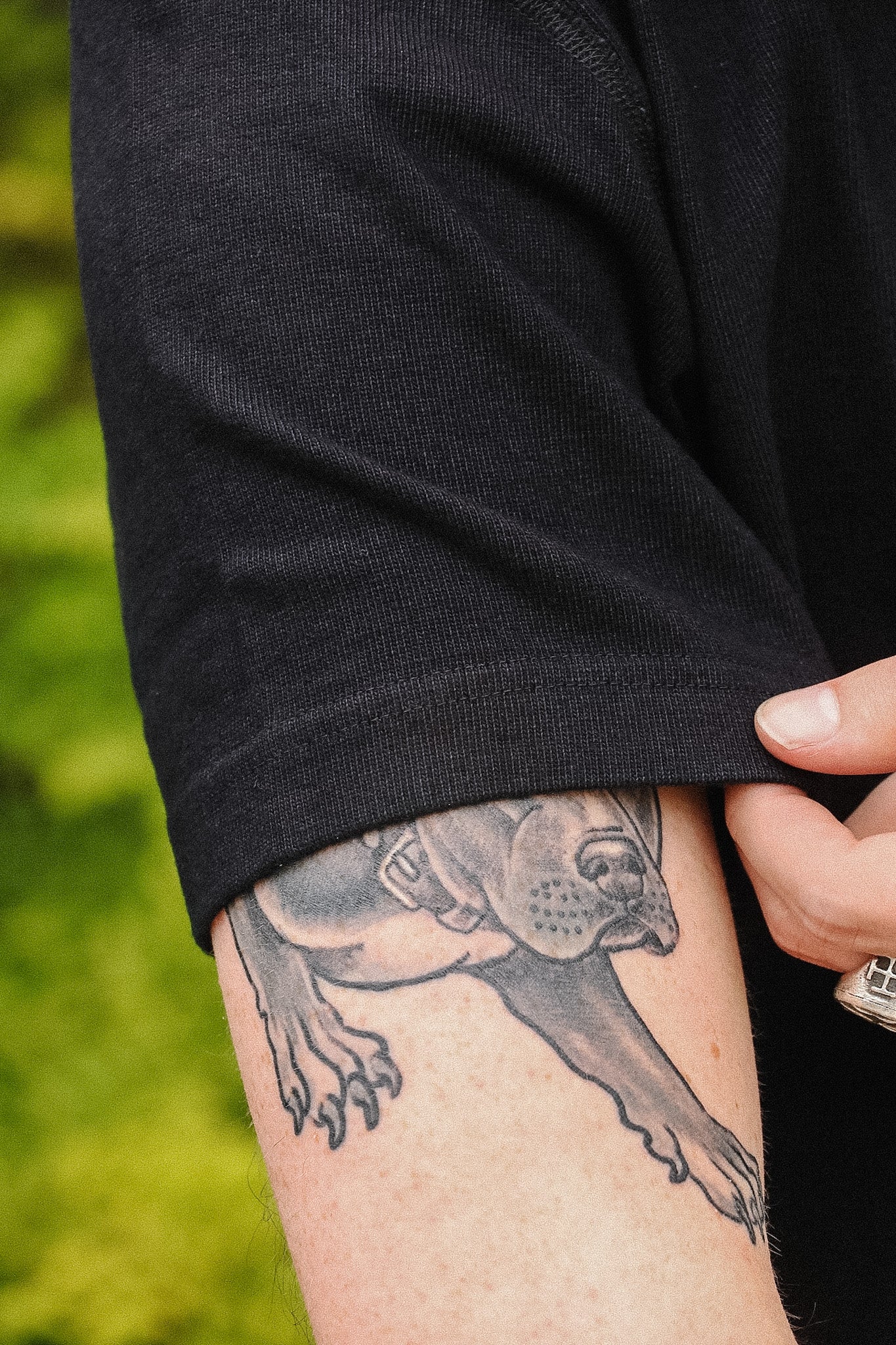A close-up of a person's upper arm wearing a black short-sleeve shirt, with a detailed black and gray tattoo of a dog visible on the skin. A hand is holding up the sleeve to show the tattoo.