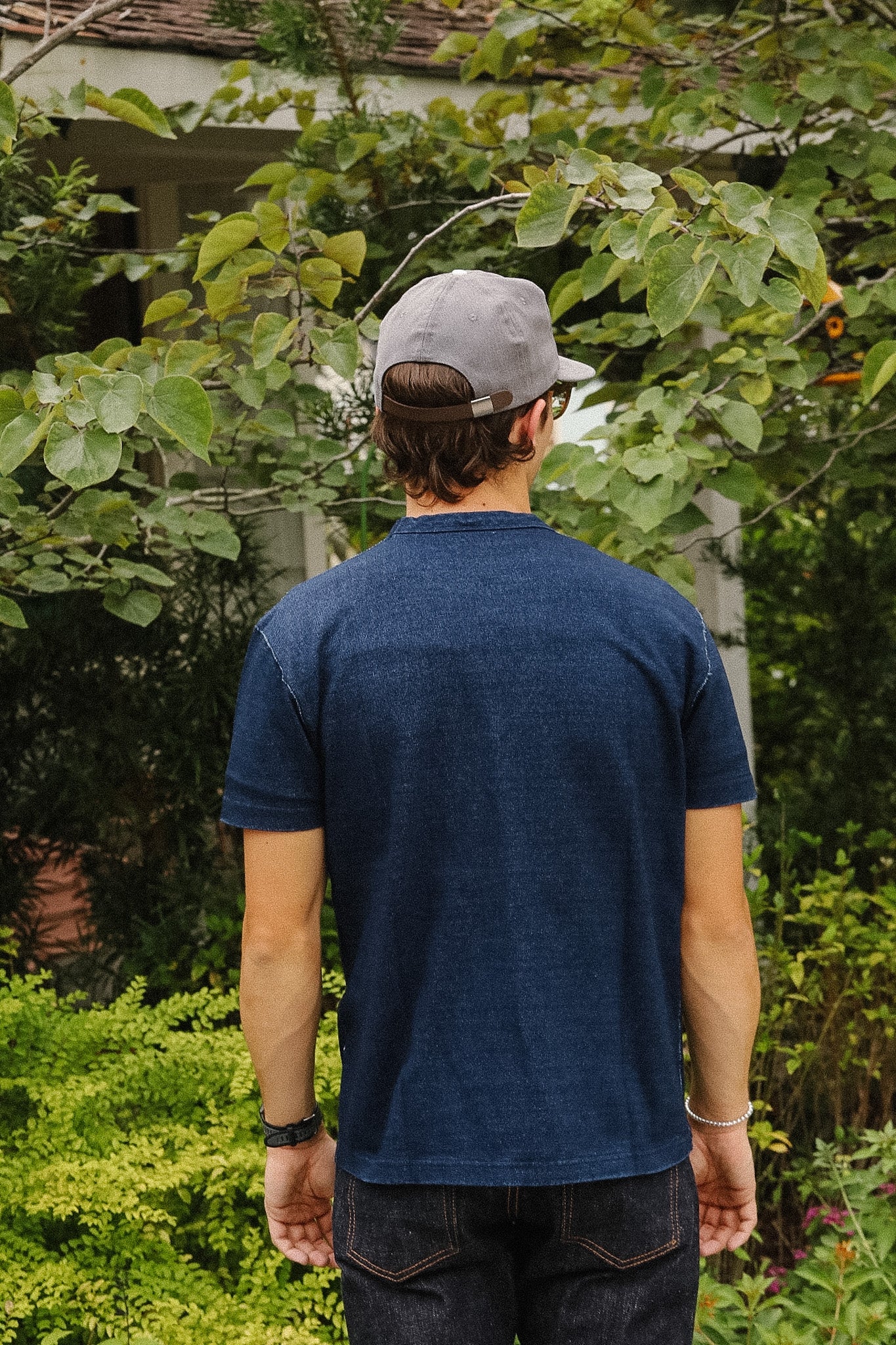 A man viewed from behind wearing a dark blue short-sleeve t-shirt, a gray baseball cap, and dark jeans, standing outdoors in front of lush green foliage.