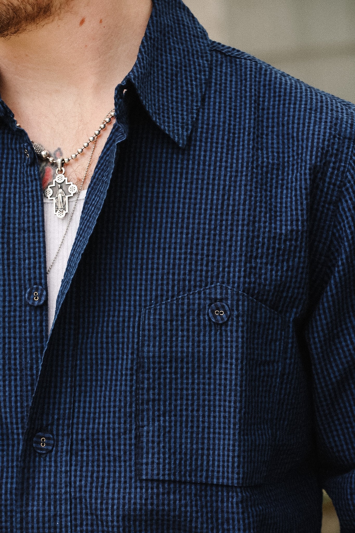 Close-up of a person wearing a dark blue and black micro-check patterned button-down shirt, layered over a white undershirt and a silver cross necklace.