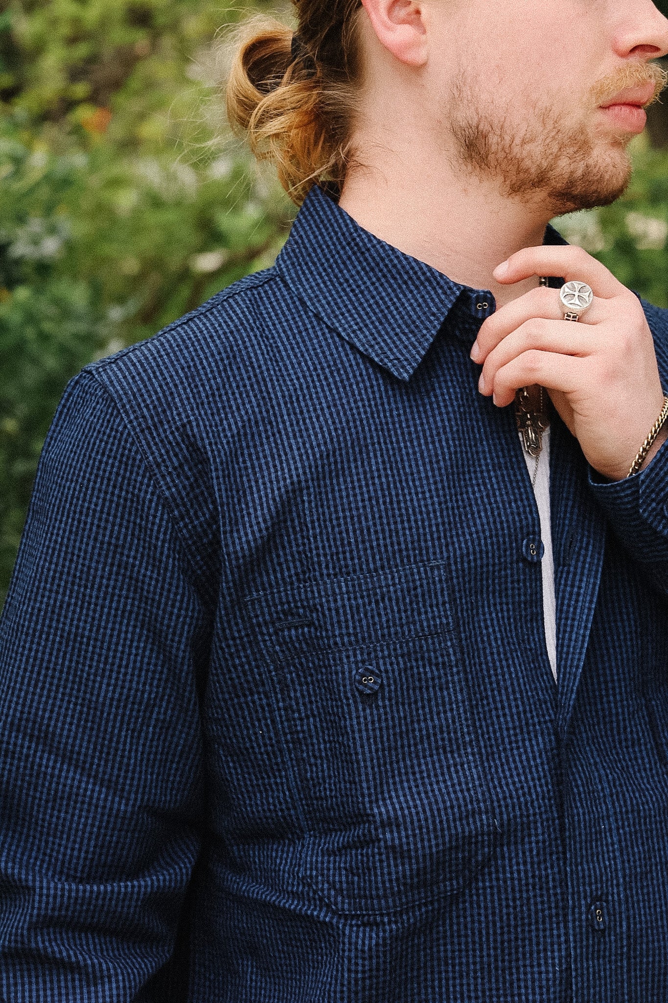 A close-up of a man wearing a dark navy blue and black micro-check patterned button-down shirt, with a white undershirt visible at the collar and a white beard.