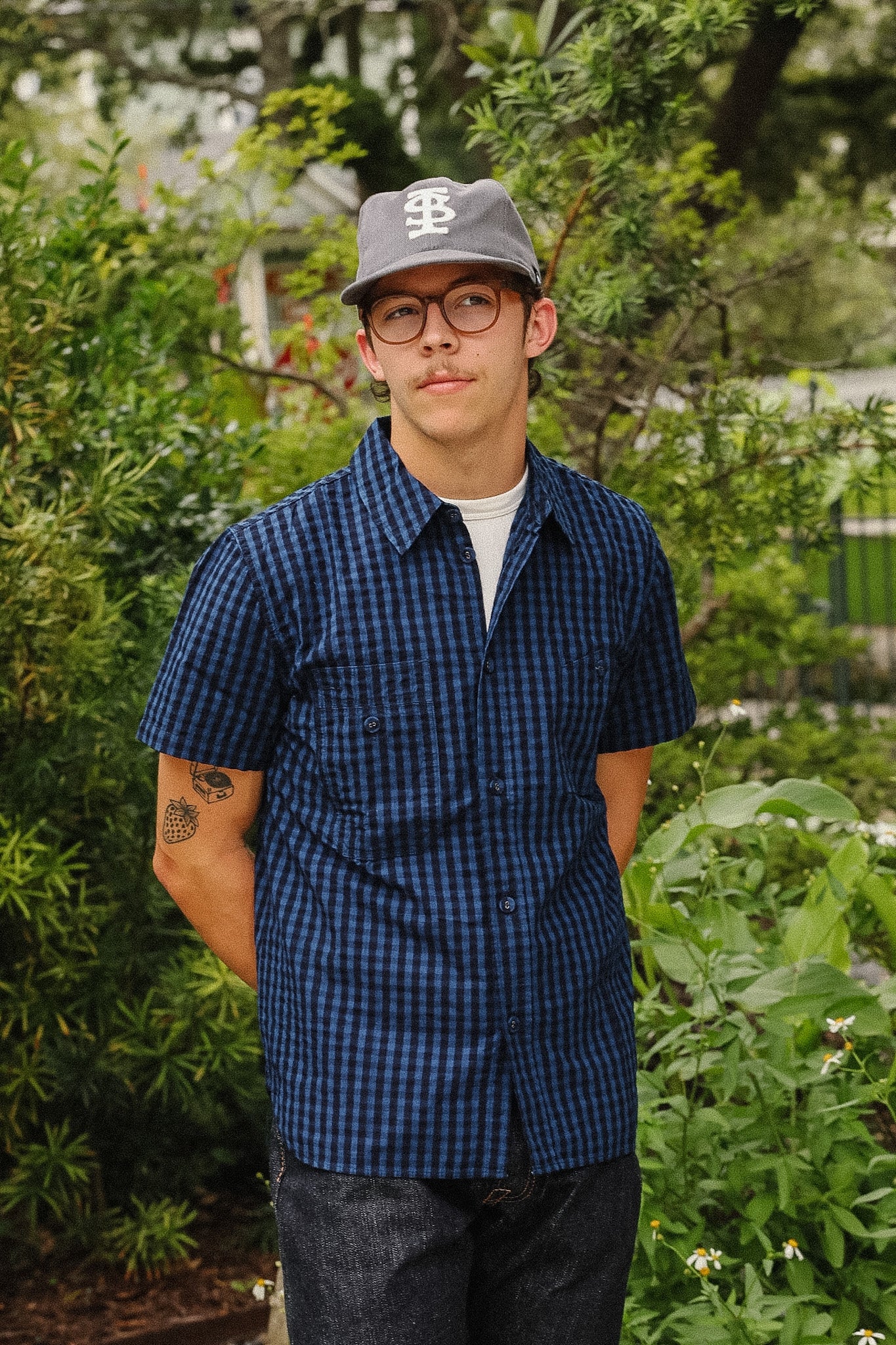 A man wearing a blue and black checkered short-sleeve button-up shirt, dark jeans, glasses, and a gray baseball cap with a white logo, standing outdoors in front of green garden foliage.
