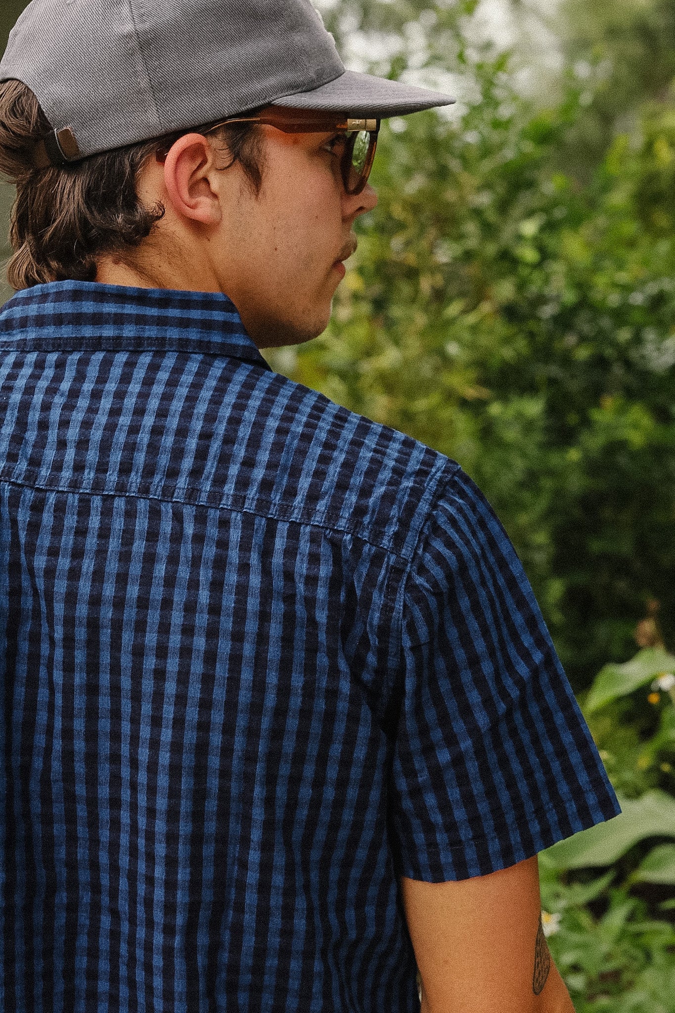 A man wearing a dark blue and black checkered short-sleeve button-up shirt, a gray baseball cap, and glasses, photographed outdoors against a blurred green foliage background.