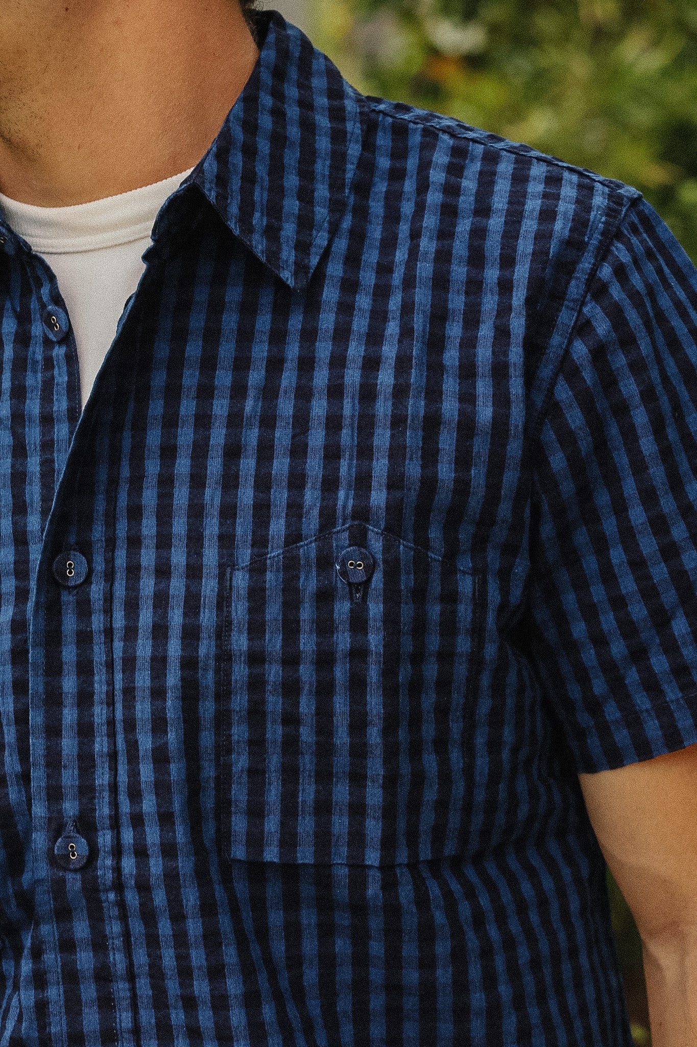 Close-up of a person wearing a short-sleeve button-up shirt with a blue and black gingham check pattern, layered over a white undershirt.
