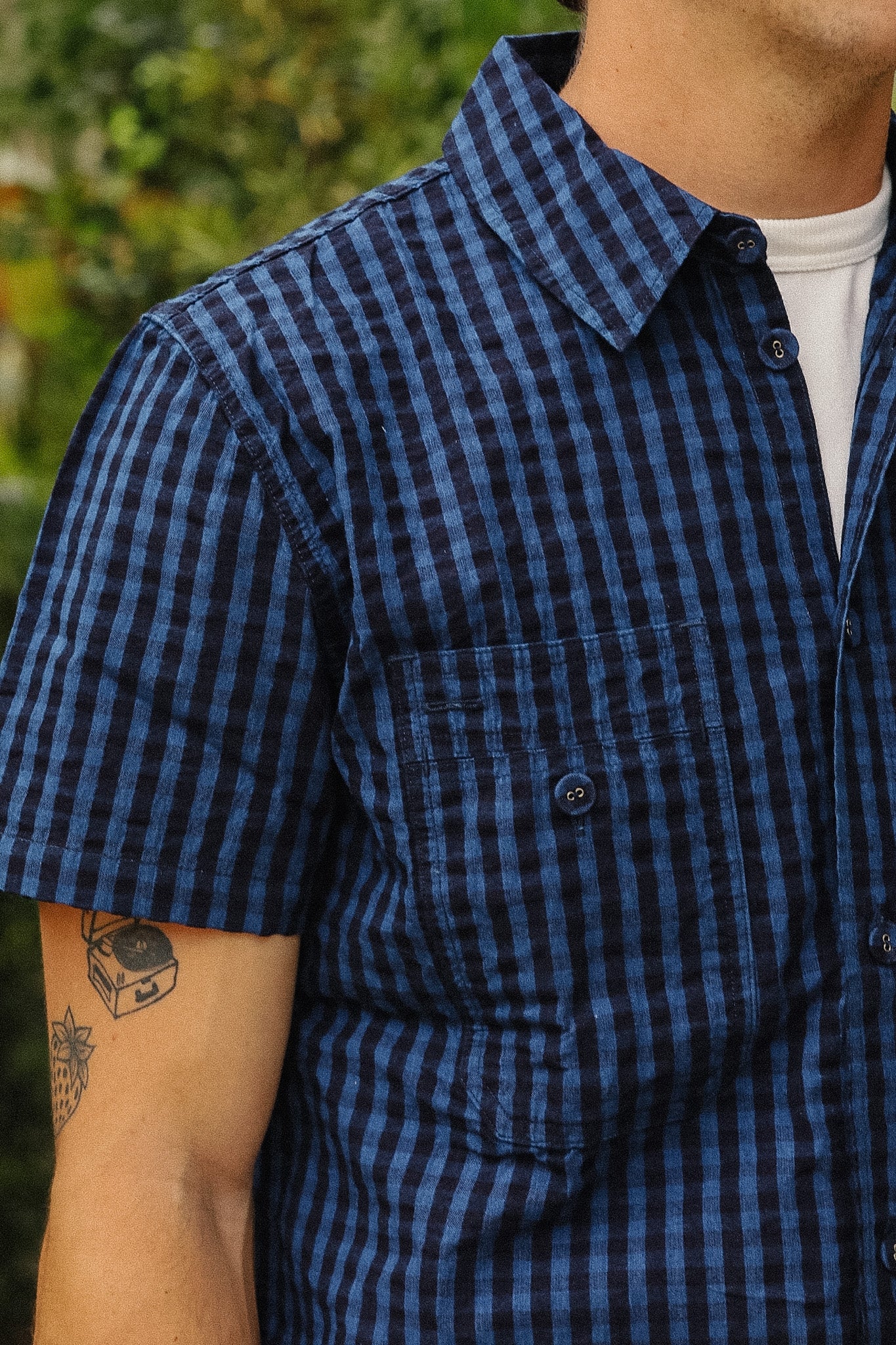 A close-up of a man wearing a short-sleeve button-up shirt with a blue and black gingham check pattern, layered over a white t-shirt. The background is a soft-focus outdoor green foliage.