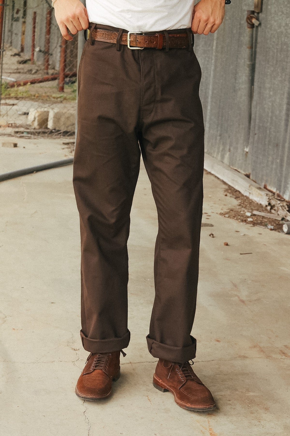 A person wearing dark brown chino pants with a brown leather belt and brown suede boots, standing outdoors against a gray concrete wall.
