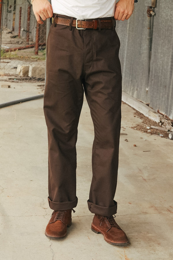 A person wearing dark brown chino pants with a brown leather belt and brown suede boots, standing outdoors against a gray concrete wall.