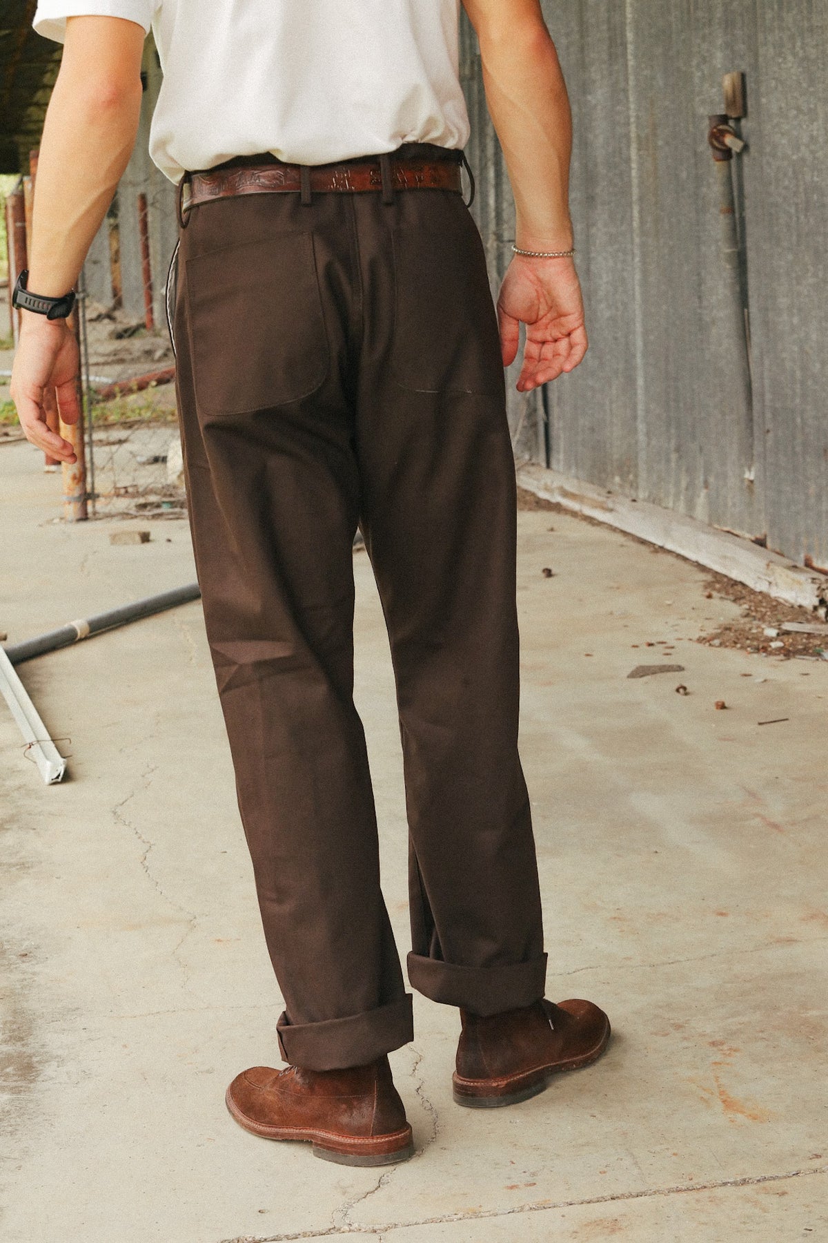 A person shown from the back wearing dark brown work pants, a white shirt, a brown belt, and brown boots in an outdoor industrial setting.