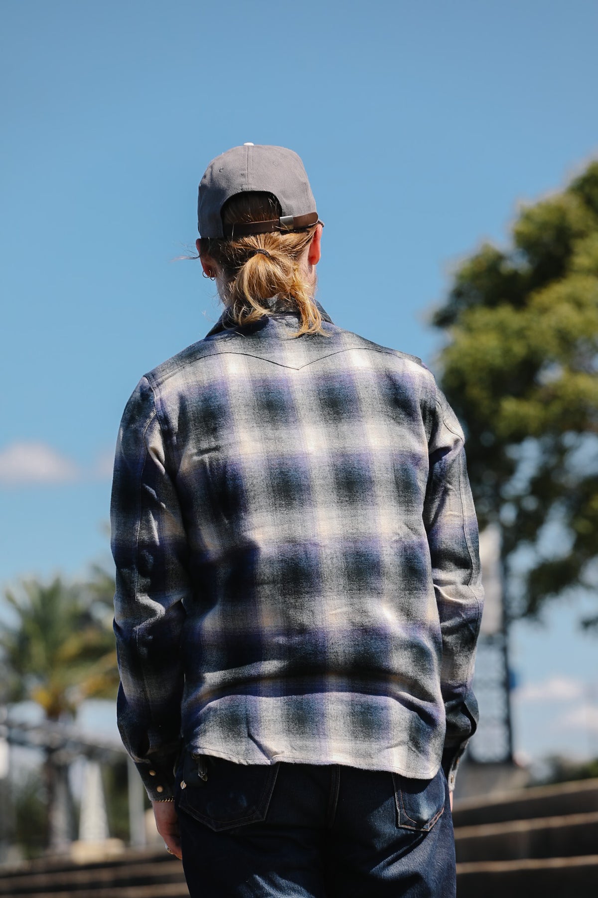 A man viewed from behind wearing a blue and gray plaid flannel shirt, dark jeans, and a gray baseball cap, standing outdoors against a blue sky and green trees.