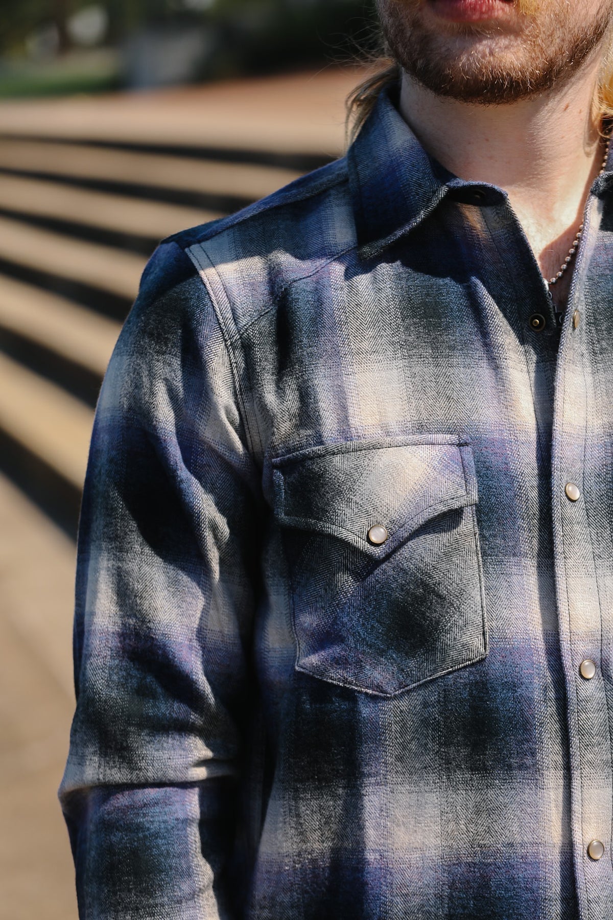 Close-up of a man wearing a blue and gray plaid flannel shirt outdoors, with sunlight casting shadows across the fabric.