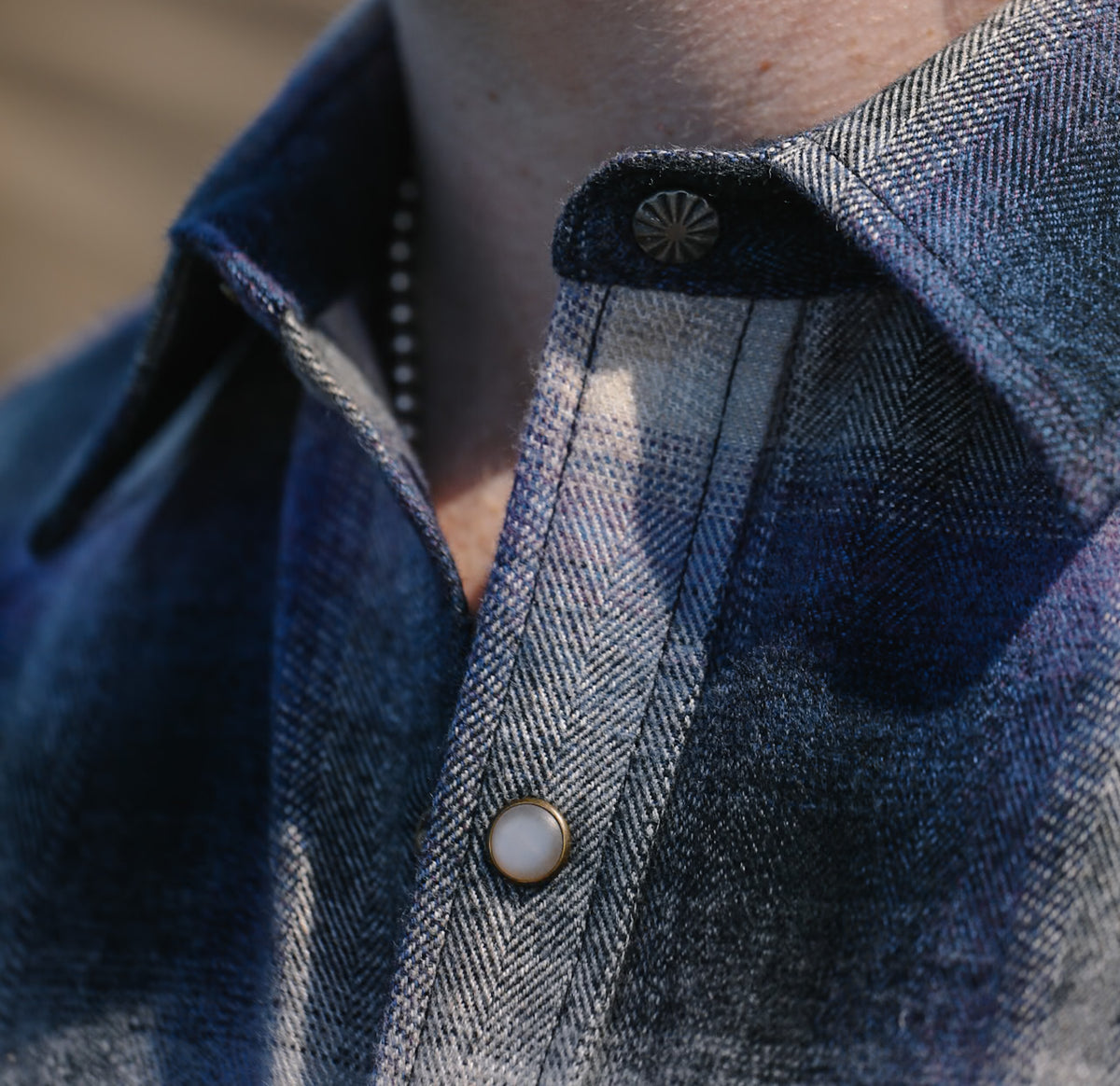 Close-up of a person wearing a textured herringbone button-down shirt in dark blue and gray tones, featuring a decorative metallic button and a pearl-like snap button.