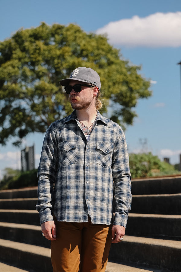 A man wearing a blue and gray plaid button-down shirt, brown pants, a gray baseball cap, and sunglasses, standing outdoors on concrete steps with a large green tree and blue sky in the background.