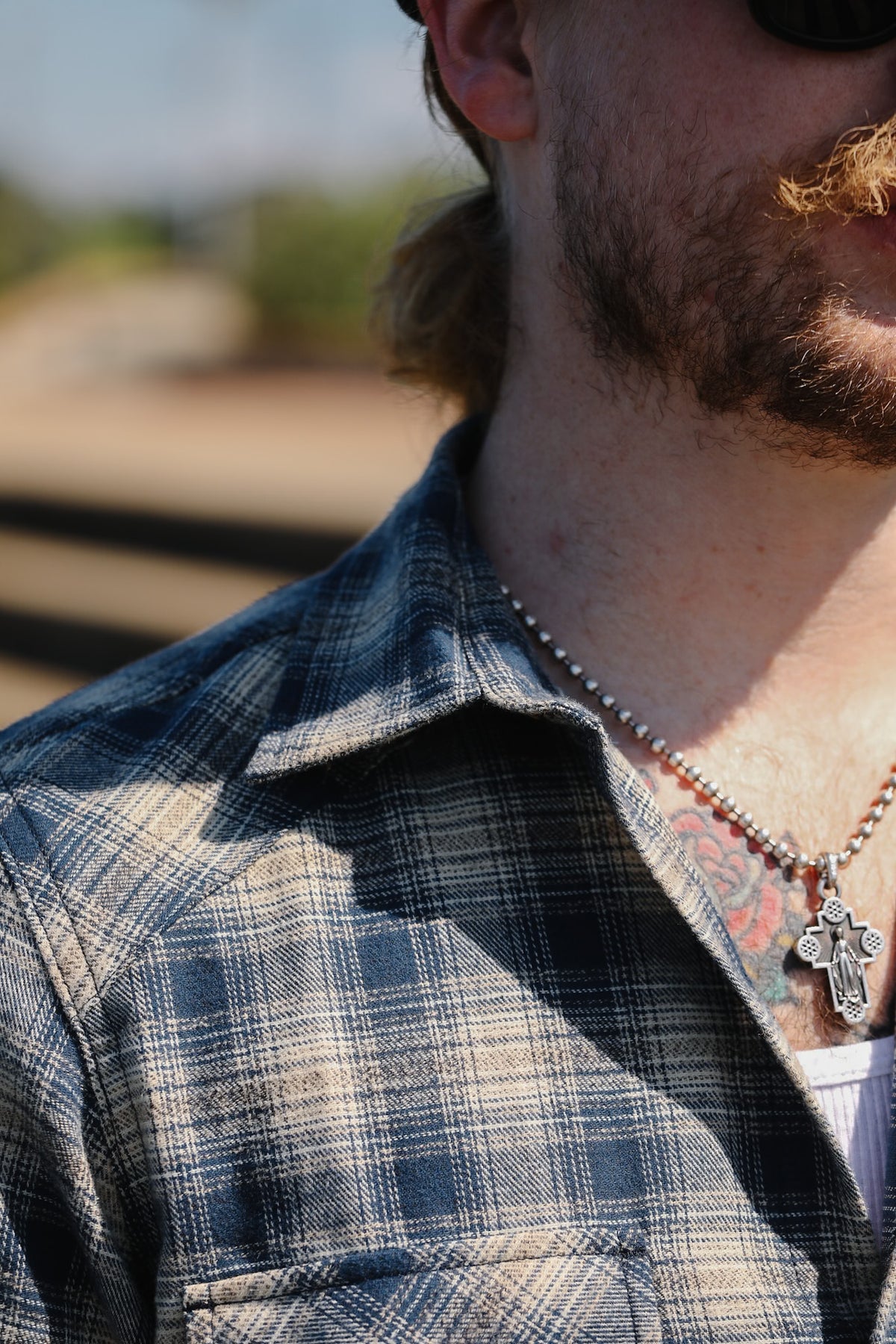 Close-up of a man wearing a blue and beige plaid button-down shirt, a silver cross pendant necklace, and sunglasses, with a blurred outdoor background.