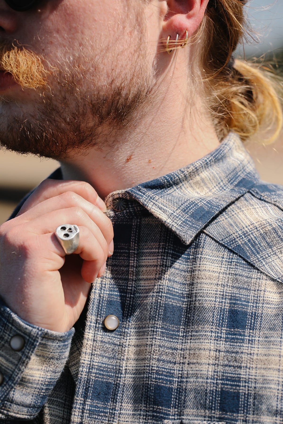 A close-up lifestyle shot of a man wearing a silver three-die rolling ring on his finger and a blue and white plaid button-down shirt.