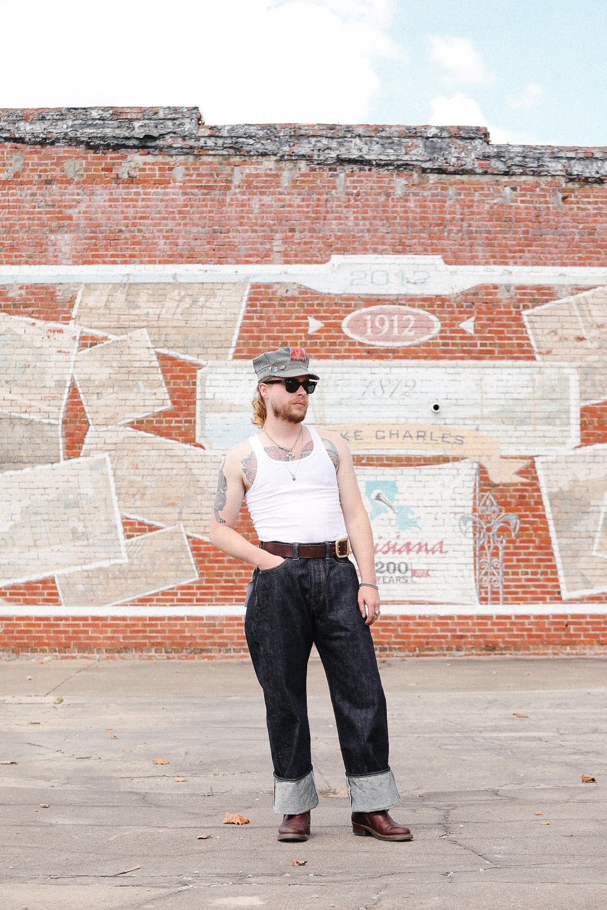 A man wearing a white tank top, dark cuffed jeans, brown boots, and a gray cap stands in front of a brick wall with a painted mural.