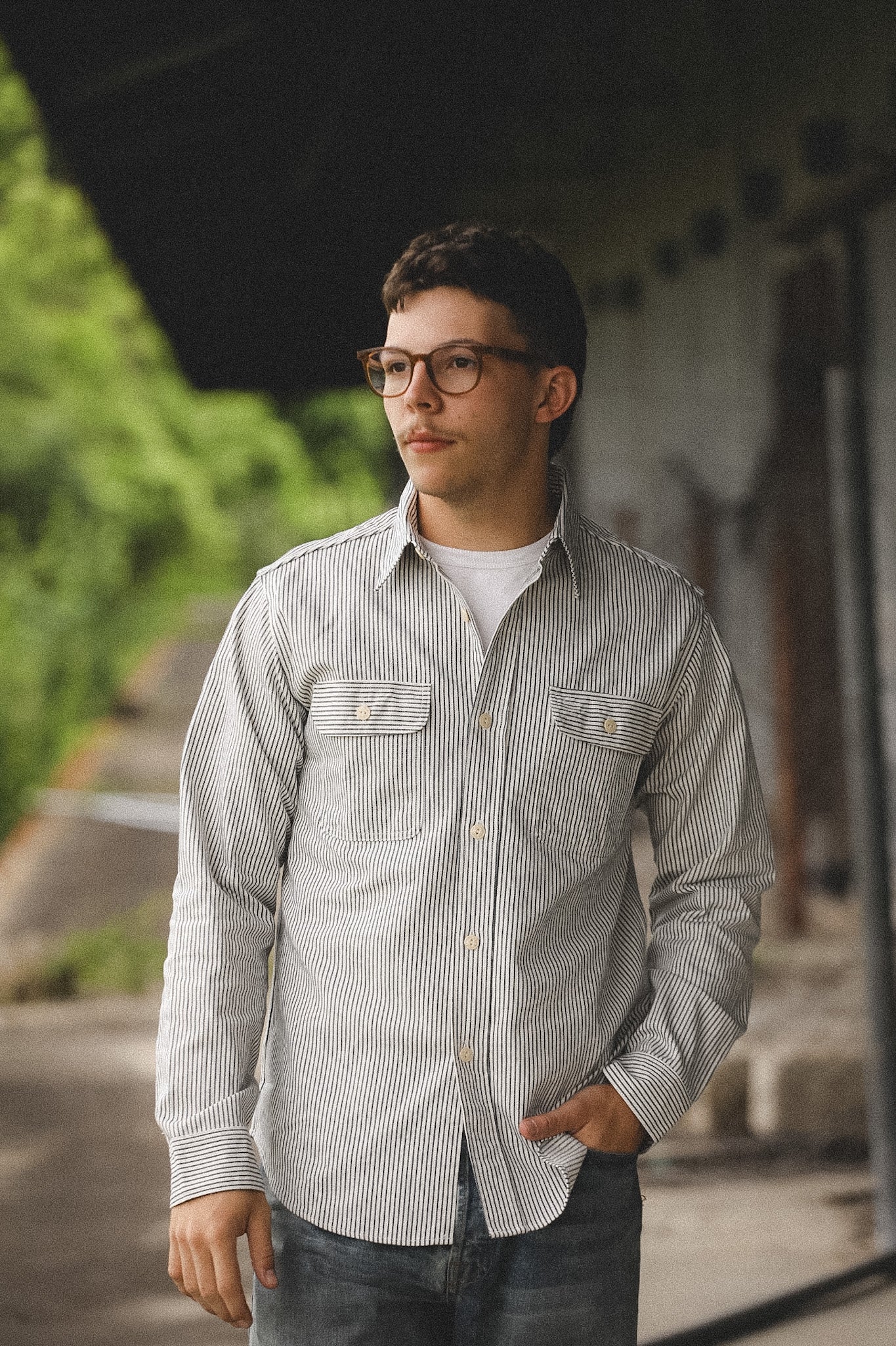 Man wearing a striped shirt and glasses standing outdoors with greenery in the background