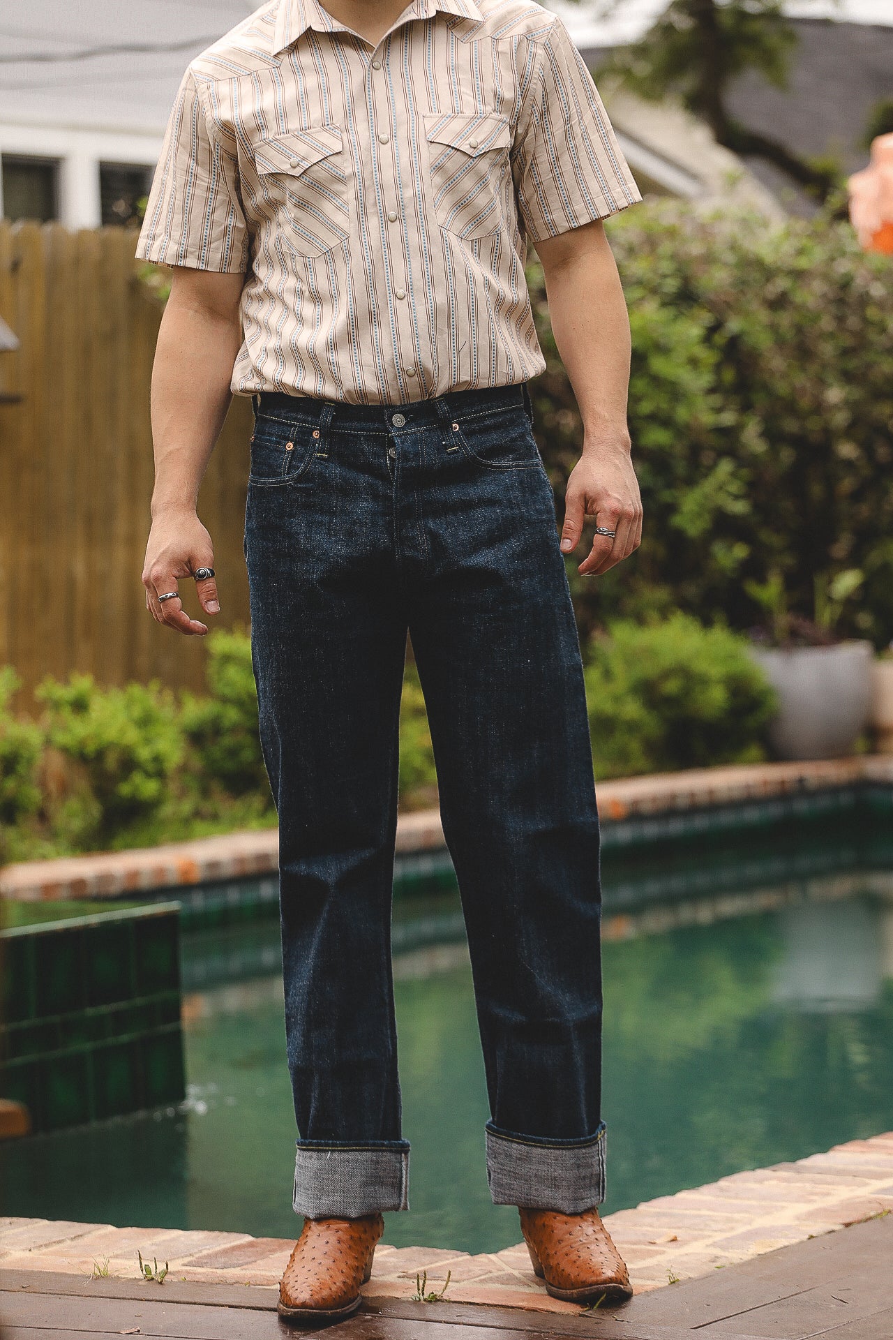 A person standing poolside wearing straight leg blue jeans and a short-sleeved striped shirt.