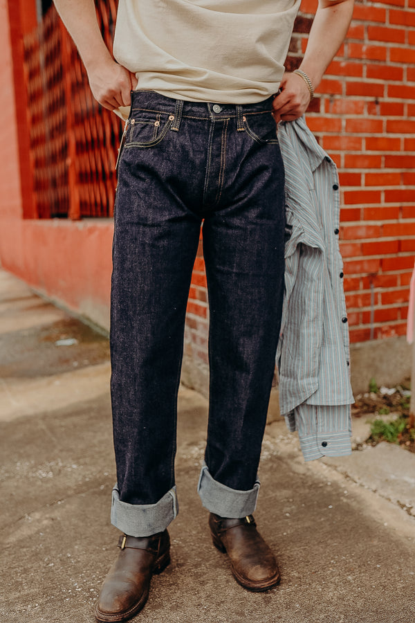 Person wearing cuffed dark denim jeans and brown boots standing on sidewalk with brick wall background