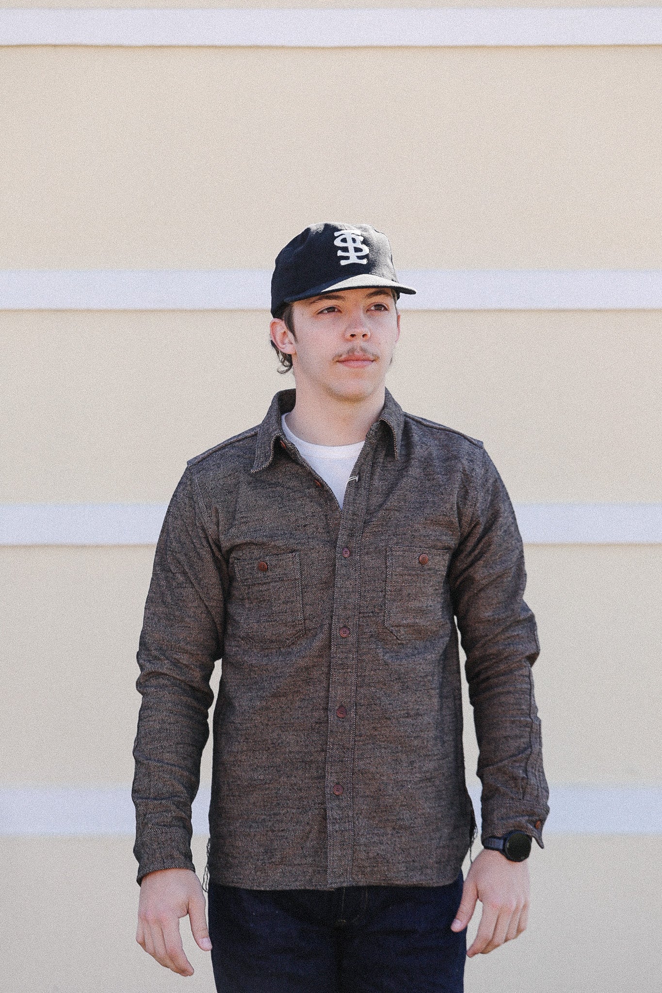 Person wearing a brown jacket and black cap against a beige wall with white stripes