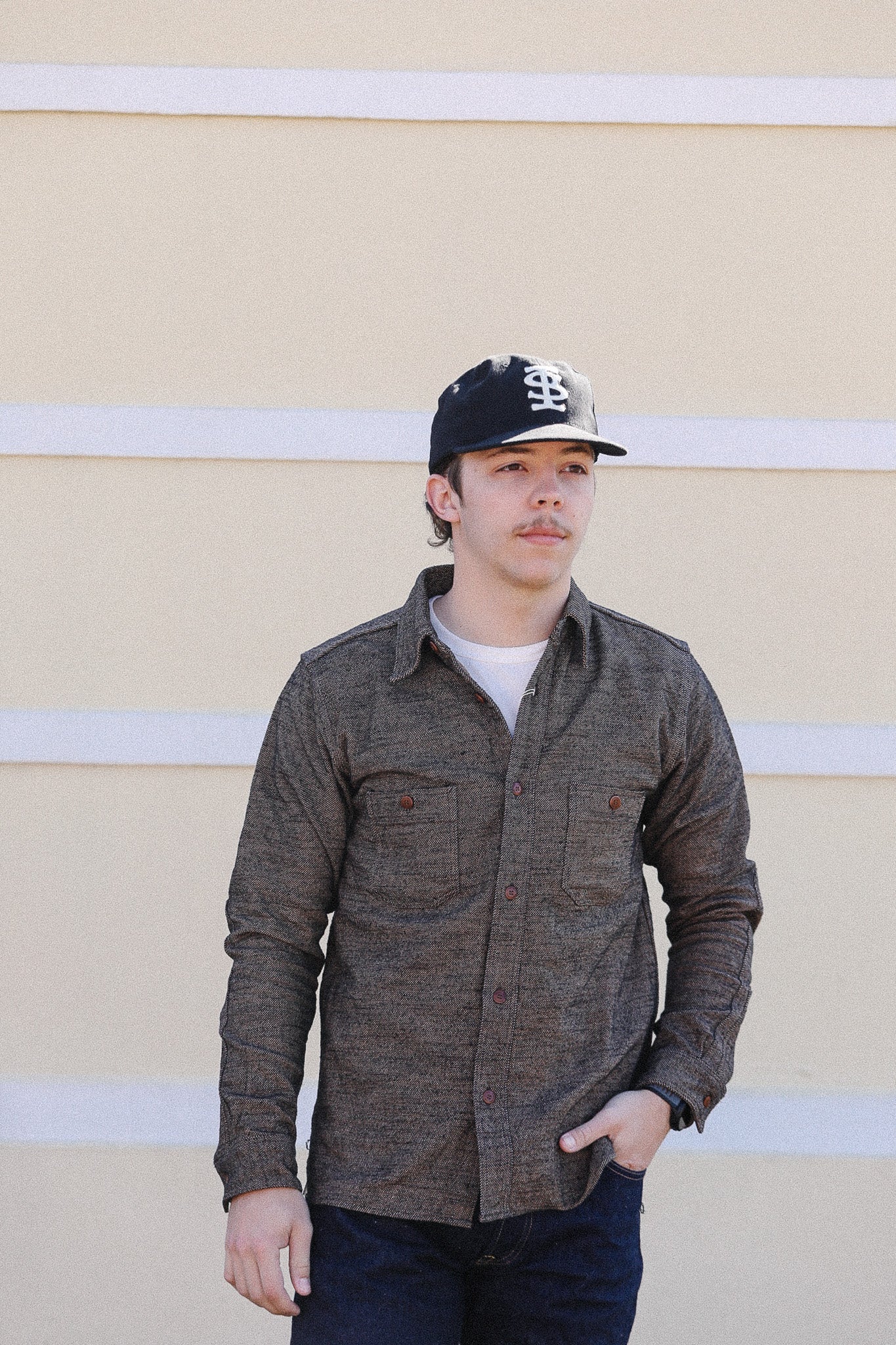 Person wearing a brown shirt and black cap against a beige wall with white stripes