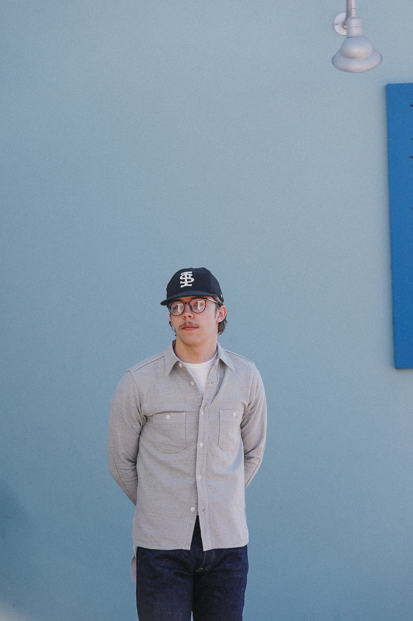 Person wearing a cap and glasses standing against a light blue wall with a blue square painting.