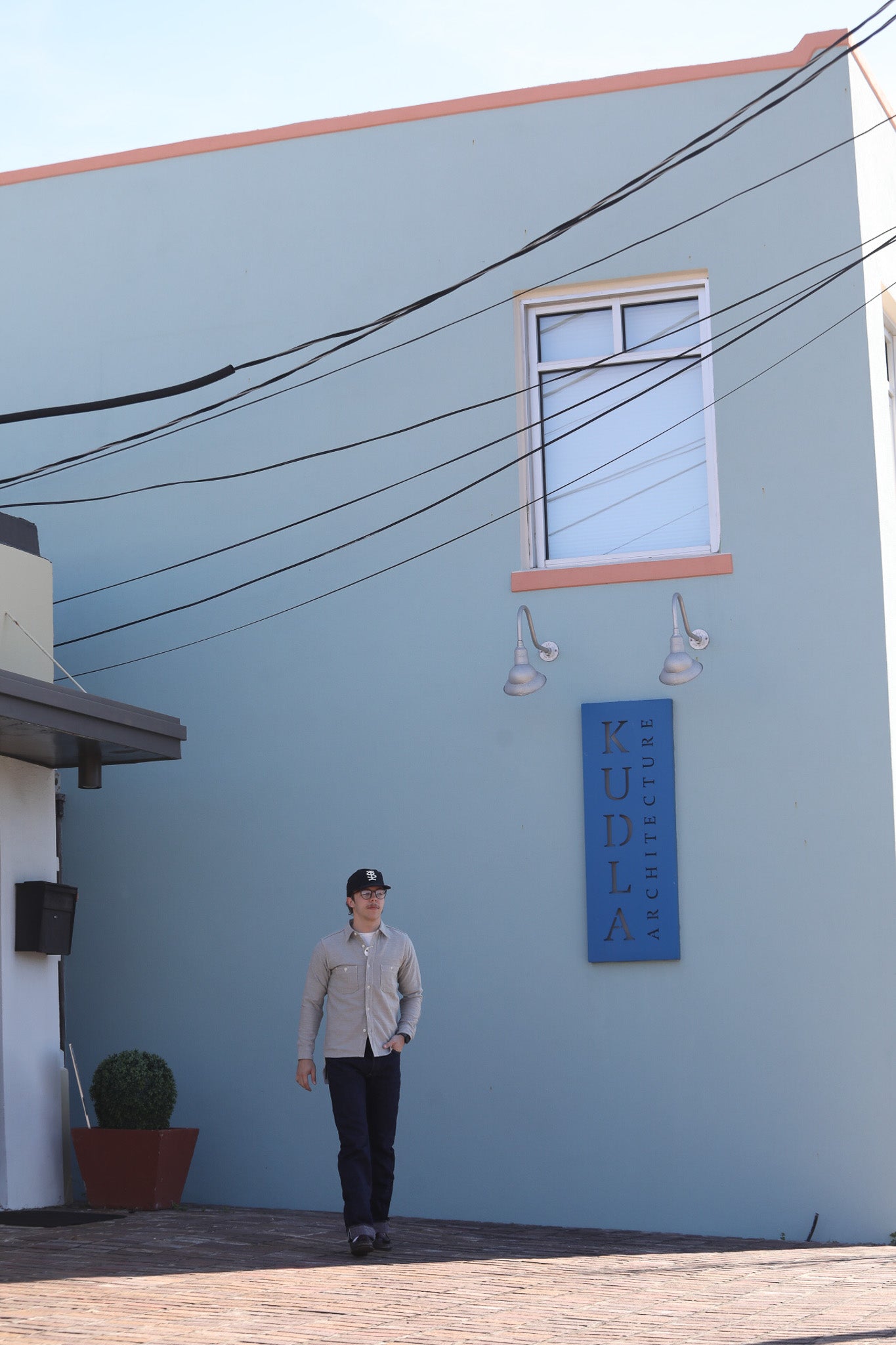 Person standing in front of a blue building with a sign, wearing a cap and light-colored shirt.
