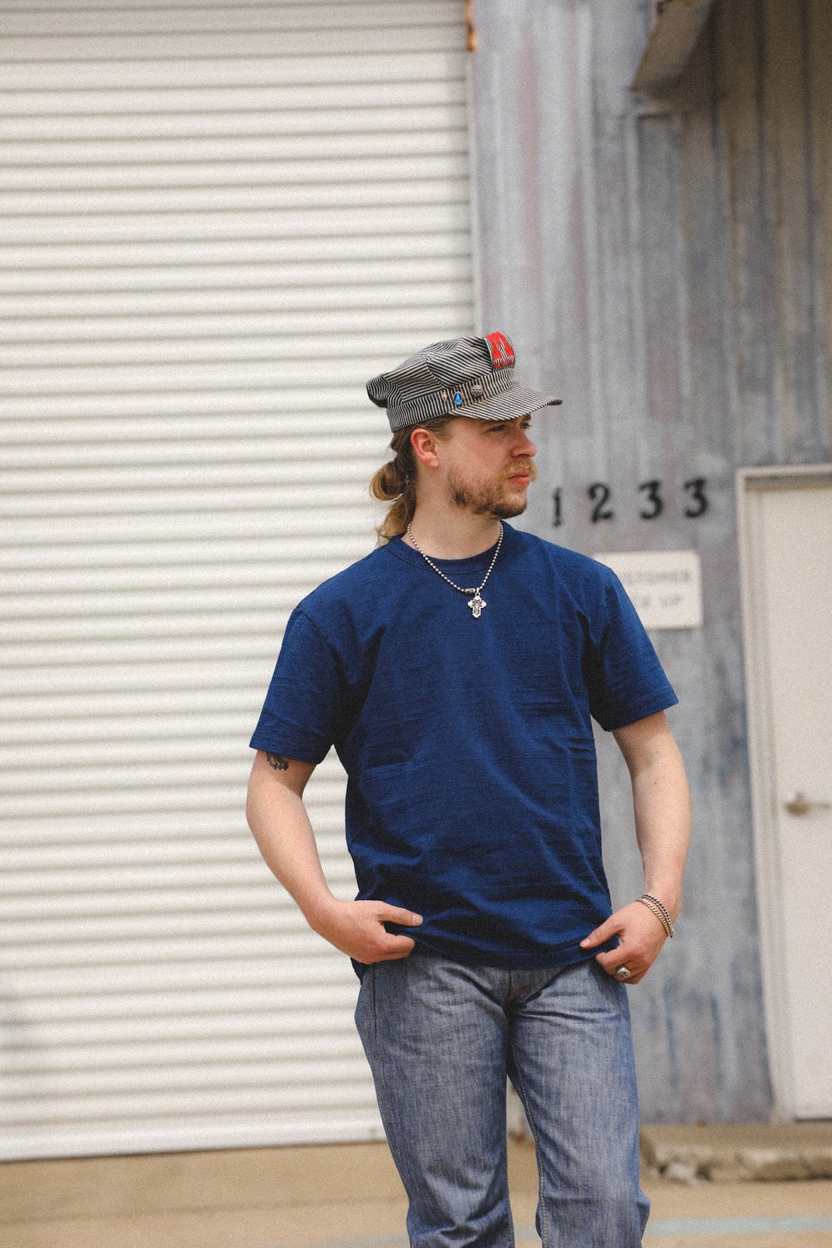 A man wearing a navy blue t-shirt, blue jeans, a gray cap, and a silver necklace stands in front of a white metal garage door and a gray building wall.