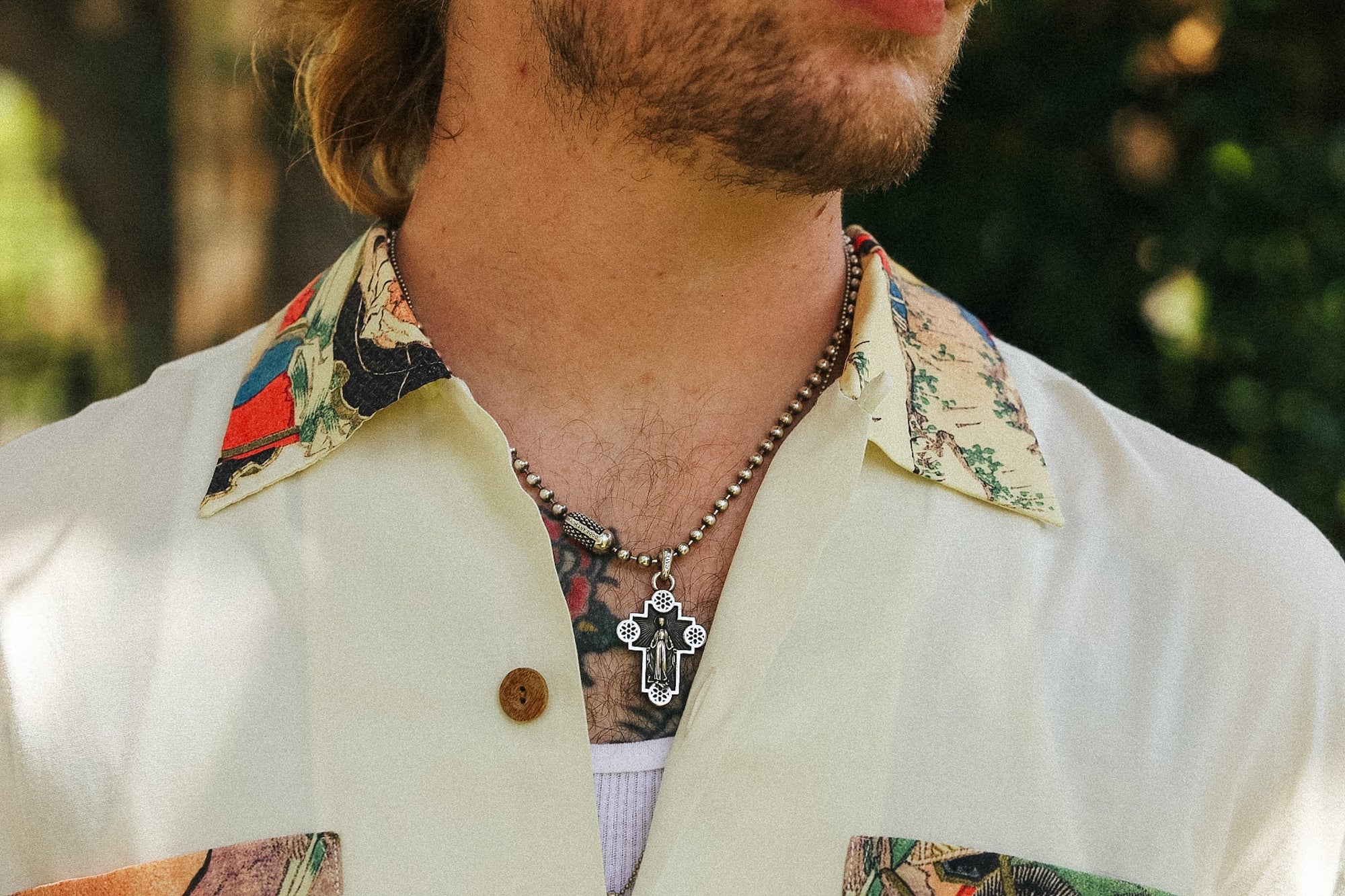A close-up lifestyle shot of a man wearing a silver-toned cross pendant necklace over a beaded chain, wearing a cream-colored shirt with a colorful patterned collar and button.