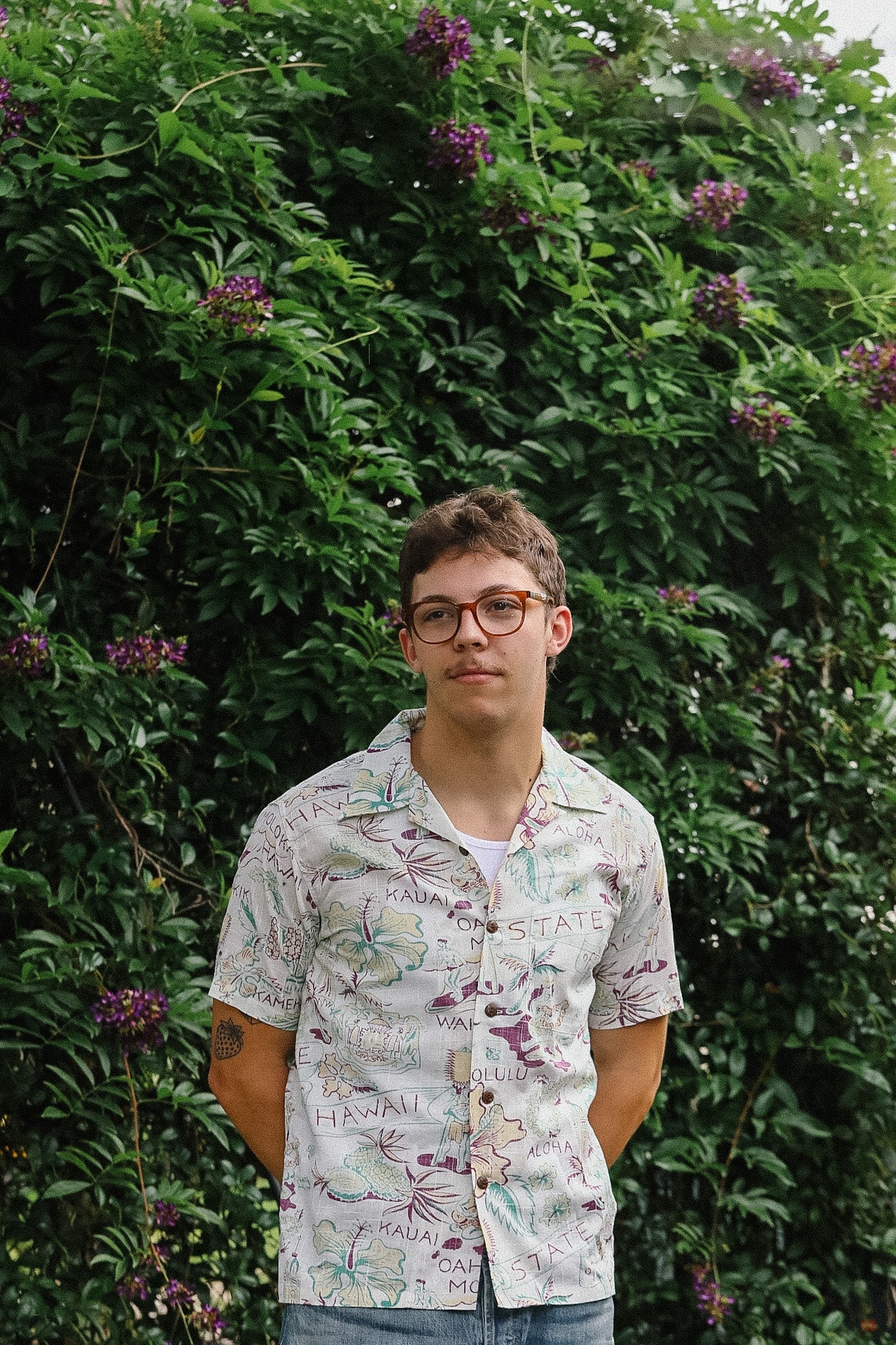 Person wearing a patterned shirt standing in front of a green bush with purple flowers