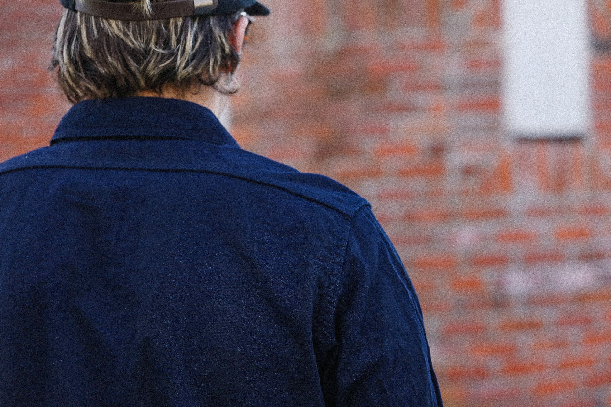 Person wearing a dark blue jacket and cap, standing in front of a brick wall.