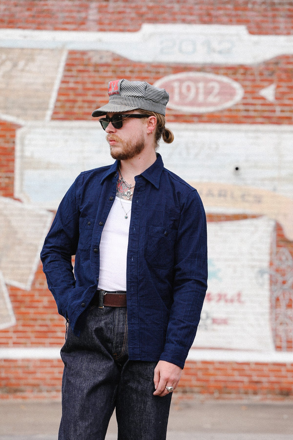 A man wearing a navy blue button-up overshirt, dark gray jeans, a white t-shirt, a gray cap, and sunglasses, standing in front of a brick wall with painted lettering.