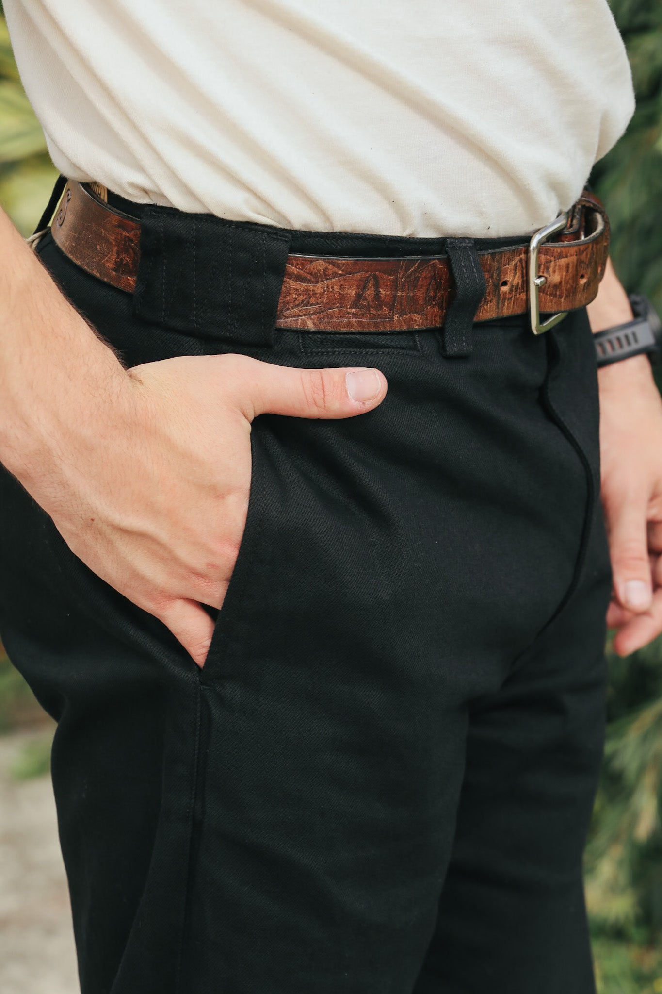 Person wearing black pants with a brown belt and beige shirt, standing against a blurred natural background.